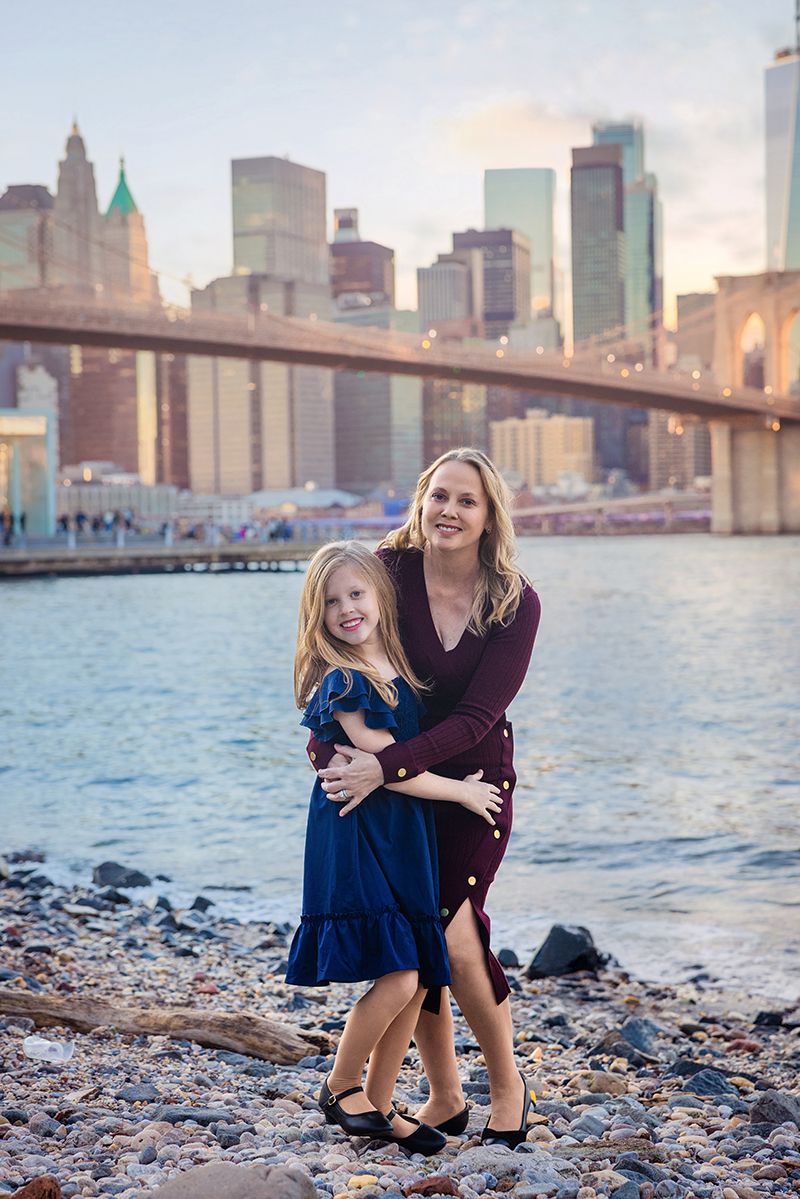 A mother and daughter pose by the Brooklyn Bridge and NYC skyline at sunset near the East River waterfront.