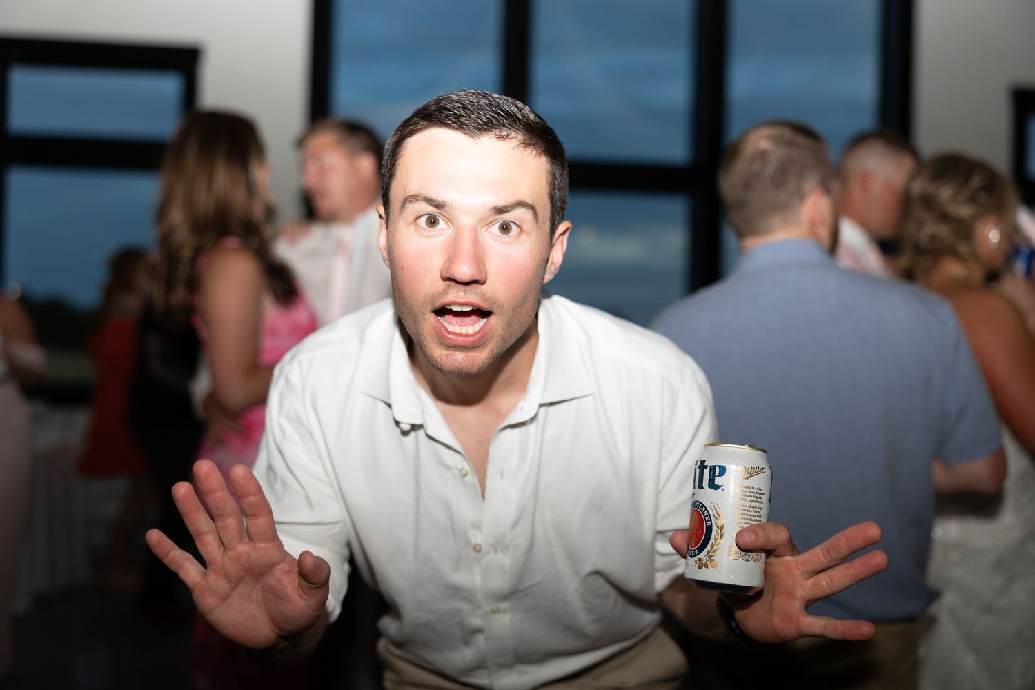 Surprised young man holding beer can with hands raised at festive wedding reception party.