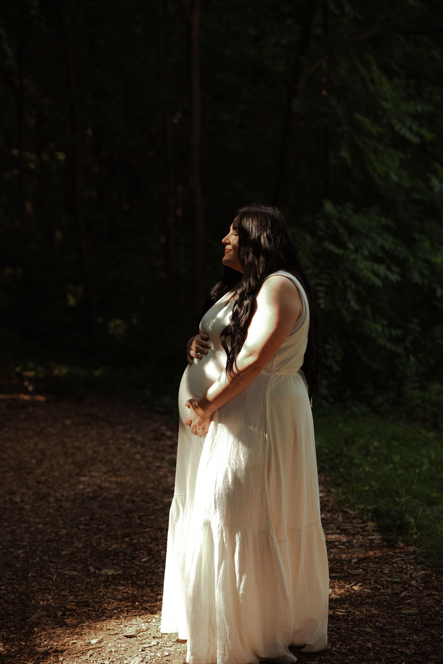 Pregnant woman in white flowing dress stands in dappled sunlight on a forest path cradling her baby bump.