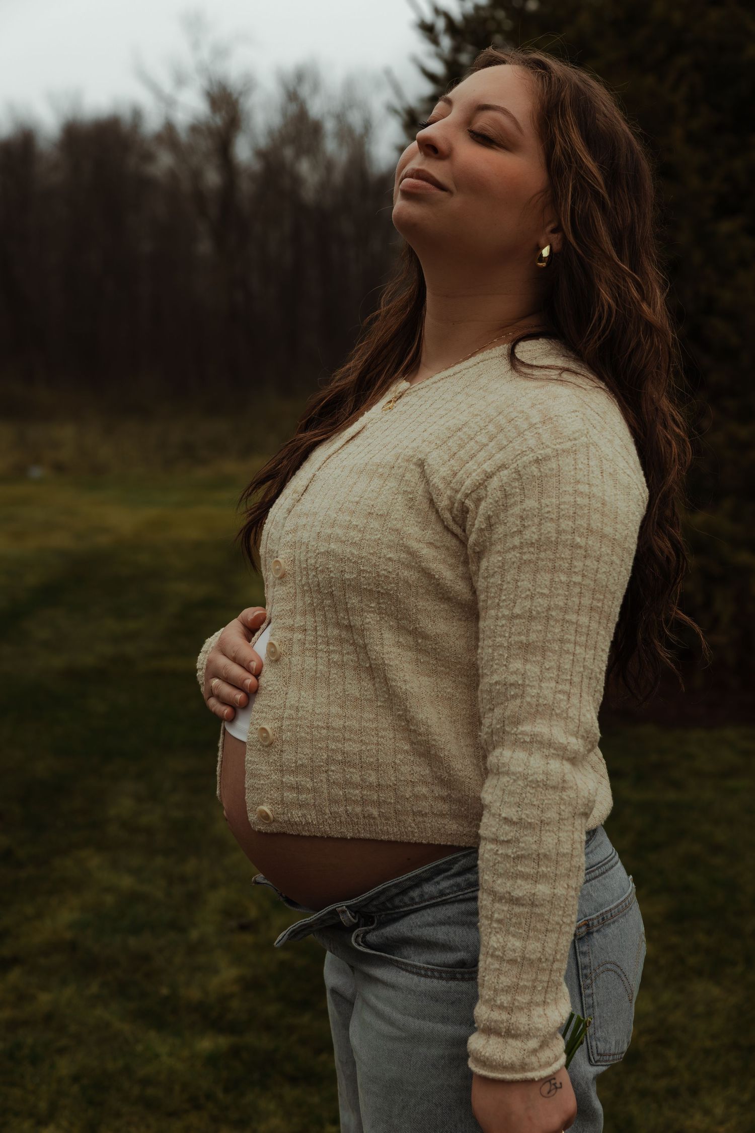 Pregnant woman in beige cardigan cradling baby bump with eyes closed outdoors.