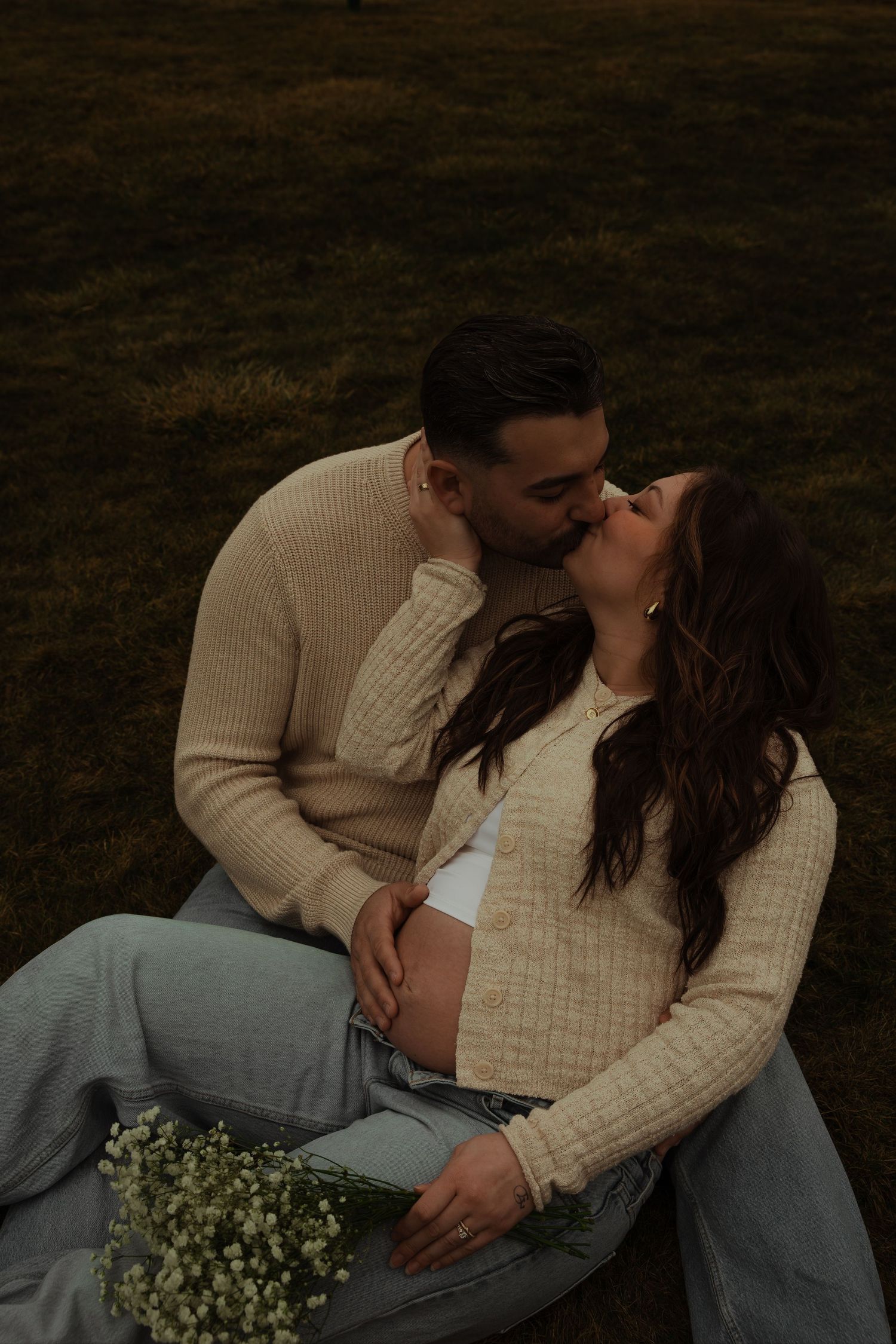 Couple sharing a kiss outdoors, woman showing pregnant belly in beige cardigan and jeans with white flowers.