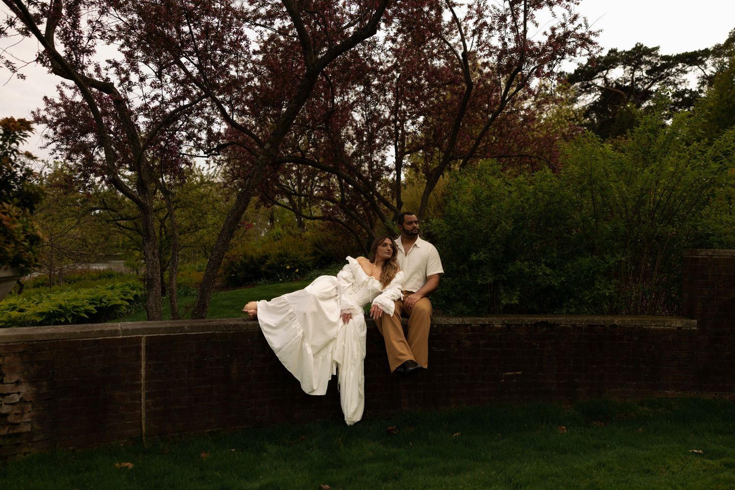 A bride and groom sit on a stone wall surrounded by lush greenery and blooming trees.