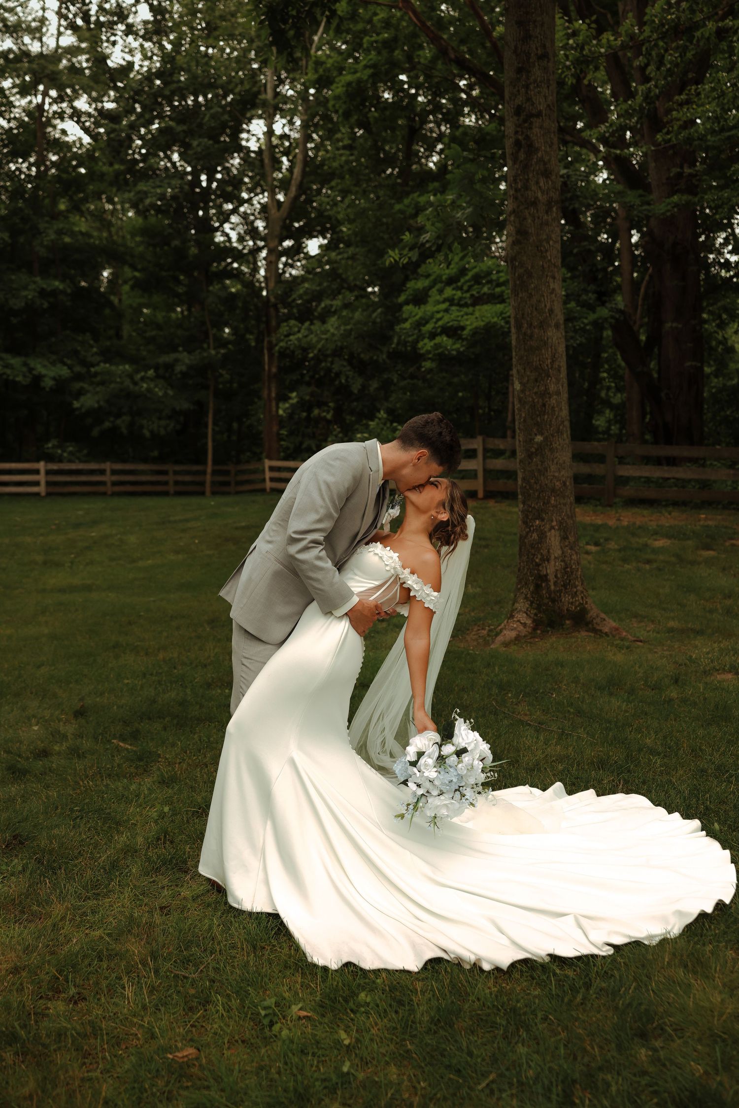 Bride and groom share a romantic dip kiss on lush green lawn surrounded by tall trees at outdoor wedding.