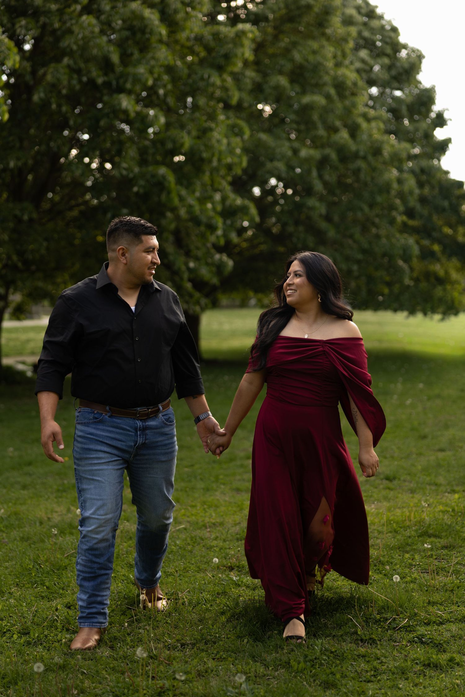 Couple holding hands walking through lush green park, woman in red off-shoulder gown.