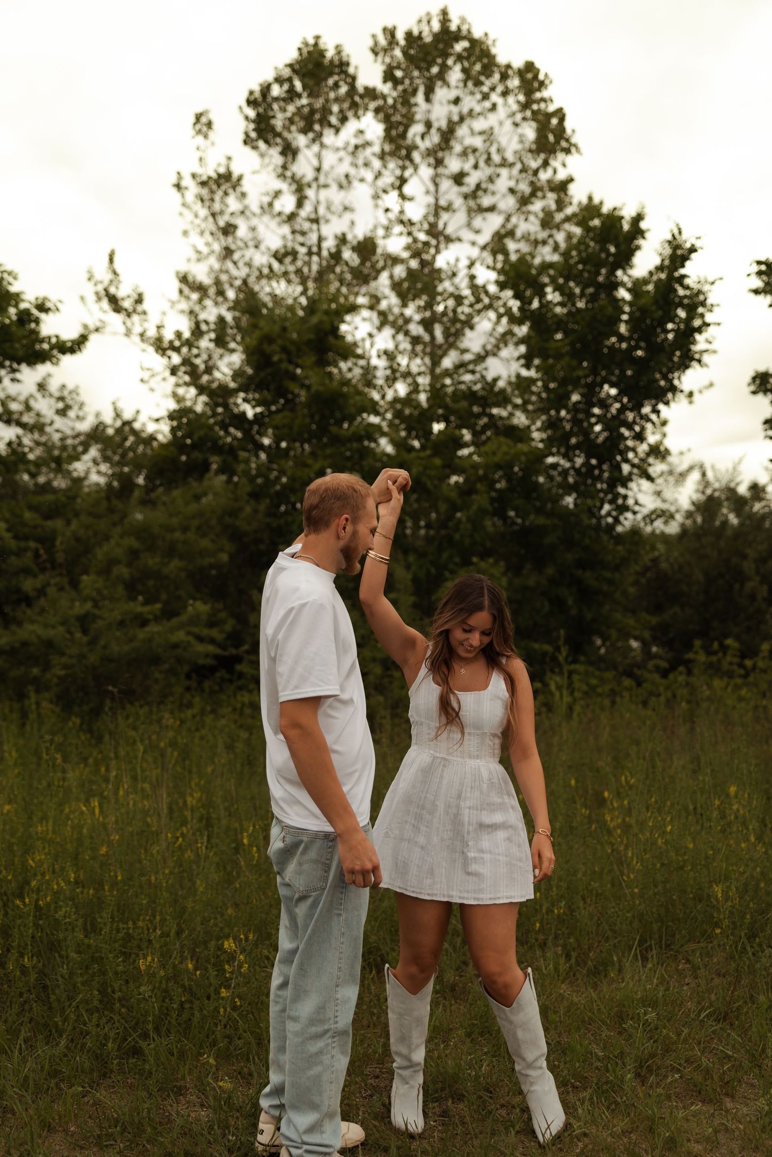 A man spins his partner in a white dress and cowboy boots in a lush green outdoor setting.