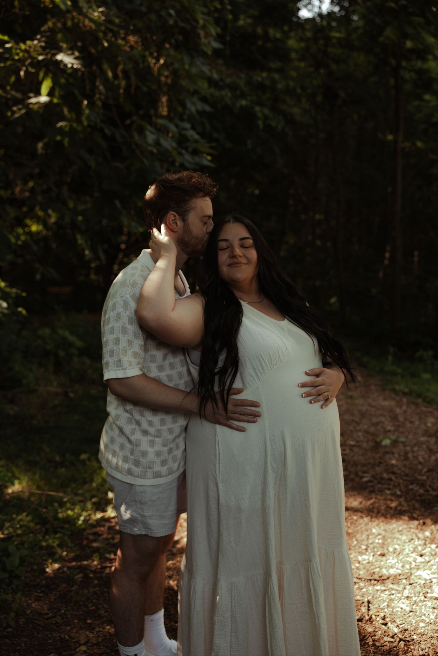 Man tenderly kisses pregnant woman's temple as she smiles, wearing a white dress on a sunlit forest trail.