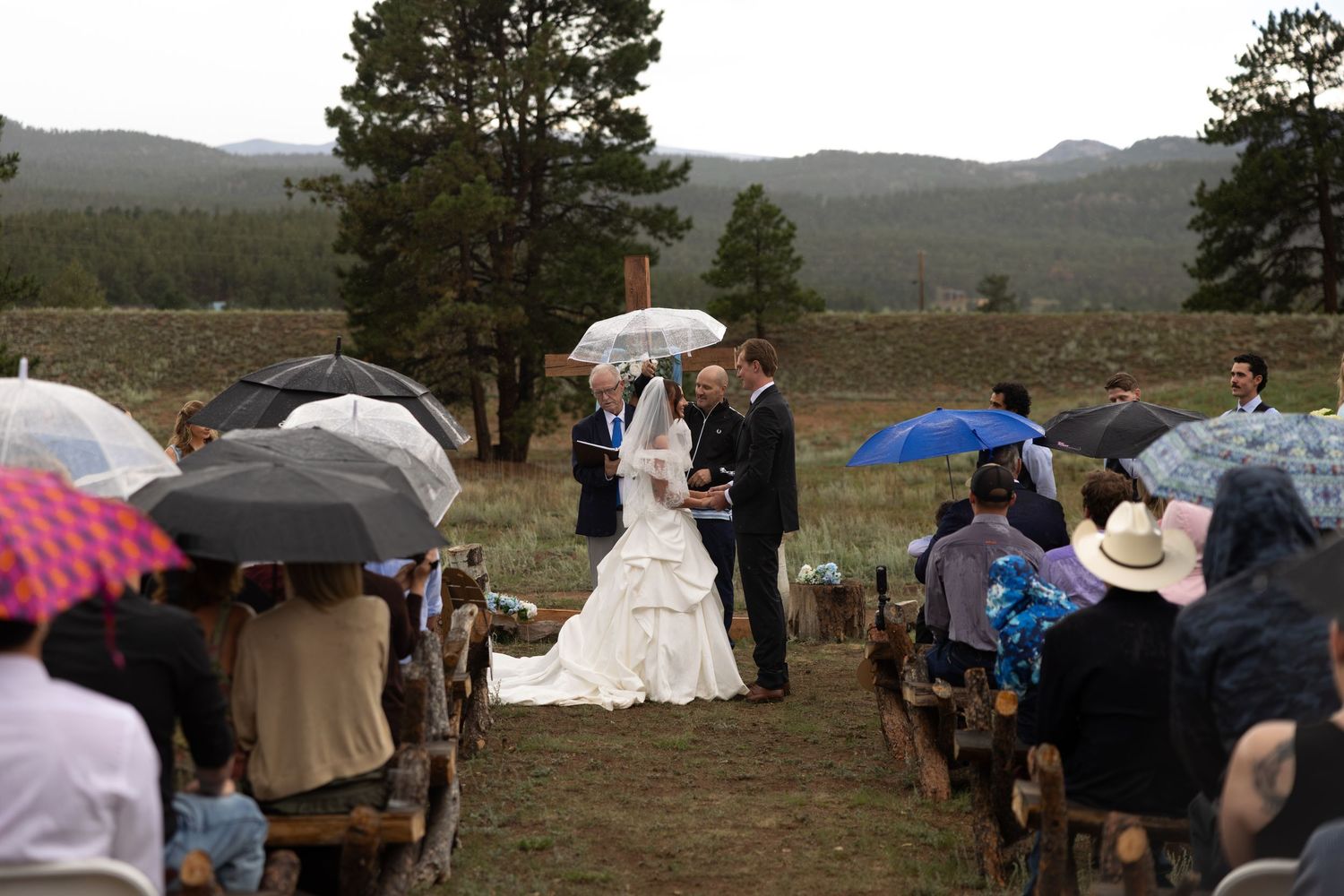 Outdoor wedding ceremony in mountain meadow with guests holding colorful umbrellas in the rain near pine forests.