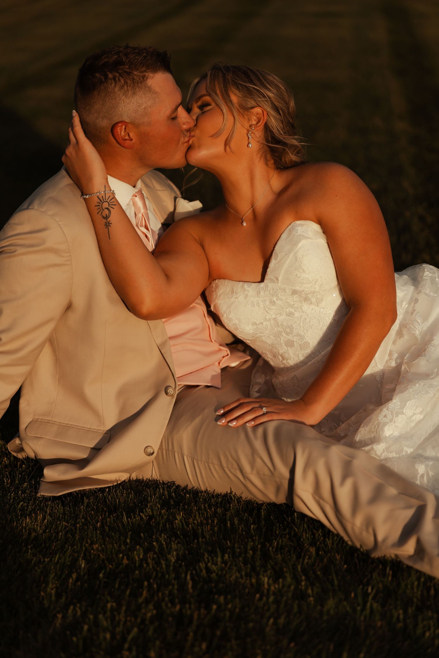 Bride and groom sharing a romantic kiss while sitting on grass at golden hour.
