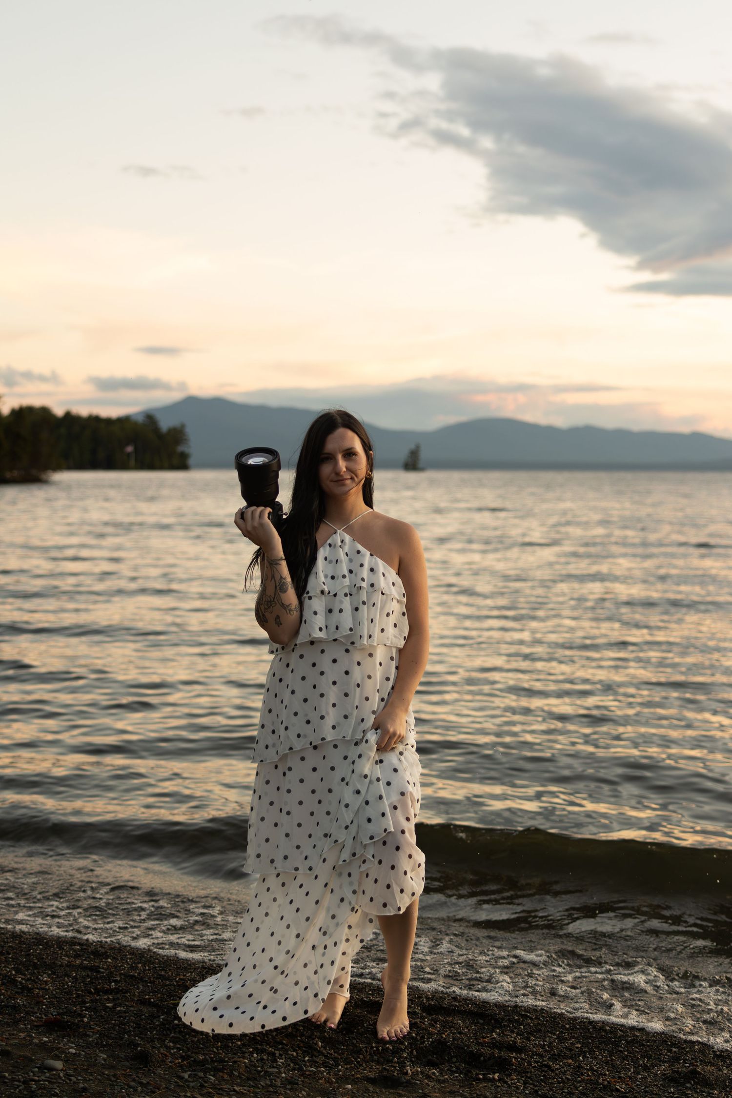 Woman in polka dot dress holding camera on rocky beach at sunset with mountains and water in background.