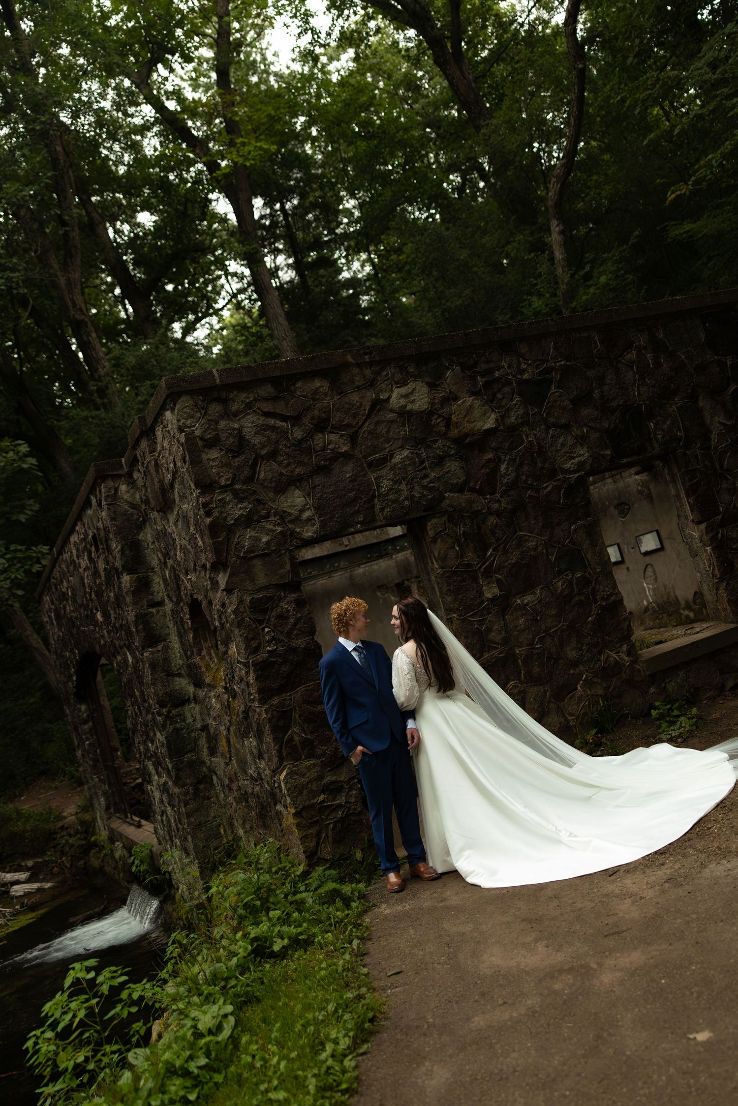 Bride in flowing white gown and veil stands with groom in navy suit before rustic stone wall surrounded by lush green trees.