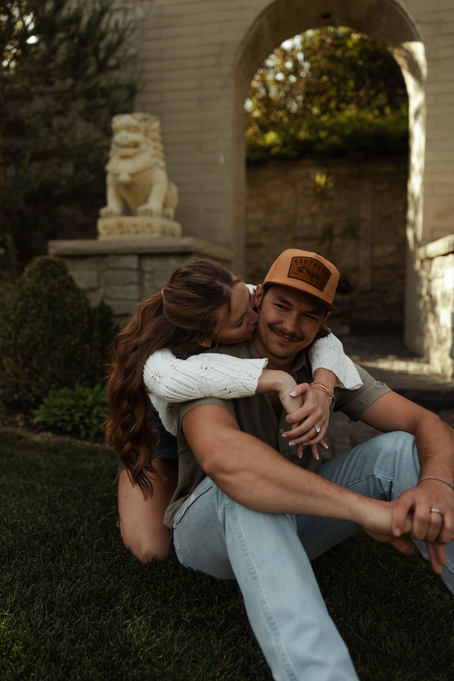 Woman kisses man's cheek as he sits on garden lawn near stone arch and lion statue.