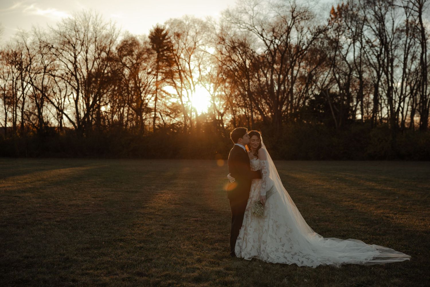Bride and groom embrace in an open field at golden sunset with bare trees in the background.