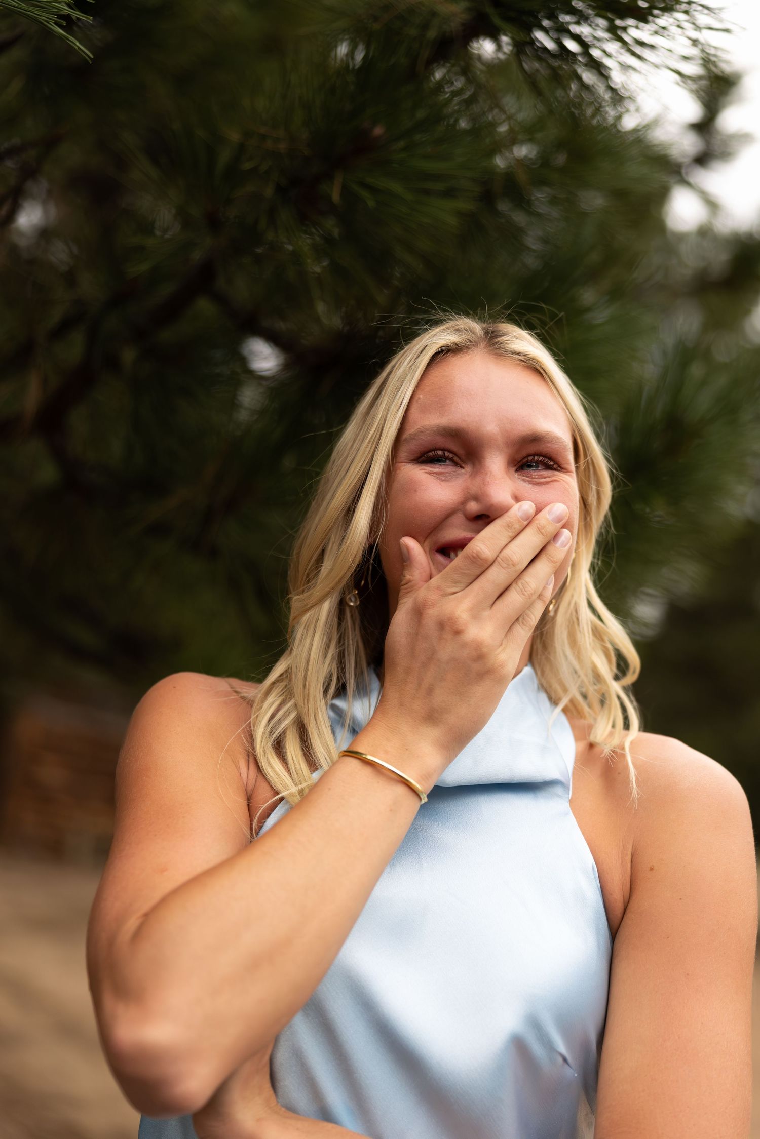 Blonde woman in light blue dress laughing with hand over mouth outdoors near pine trees.