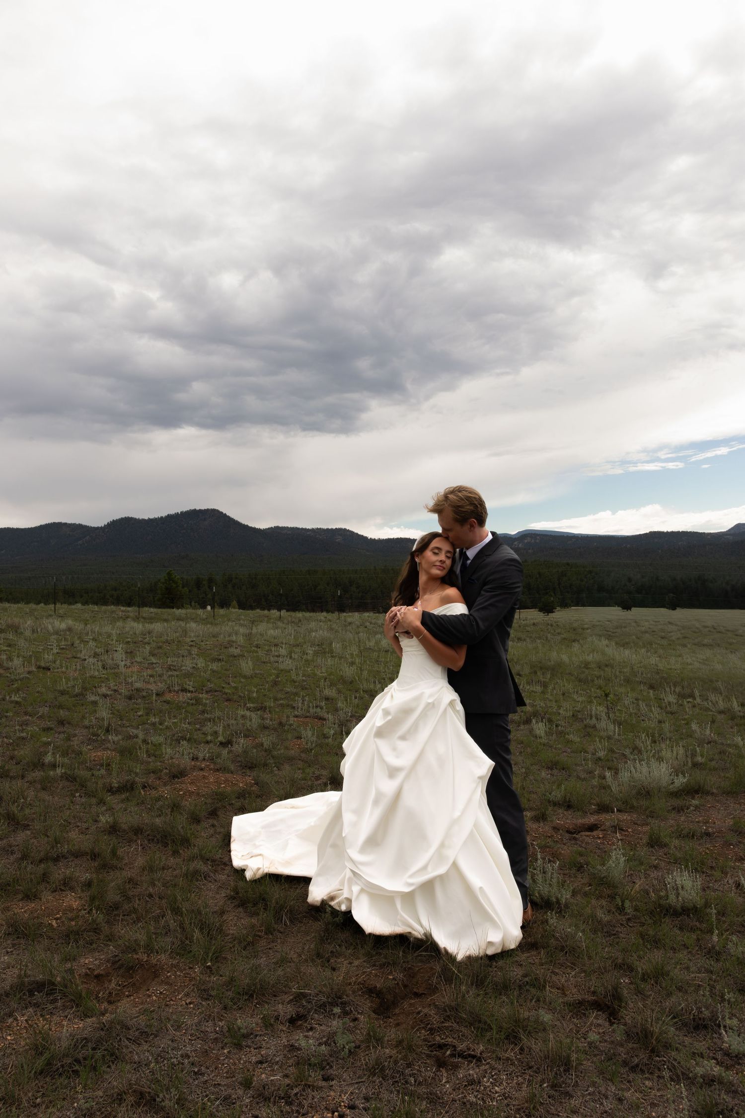 Bride and groom embrace in a scenic mountain meadow, with dramatic cloudy skies and rolling hills in the background.