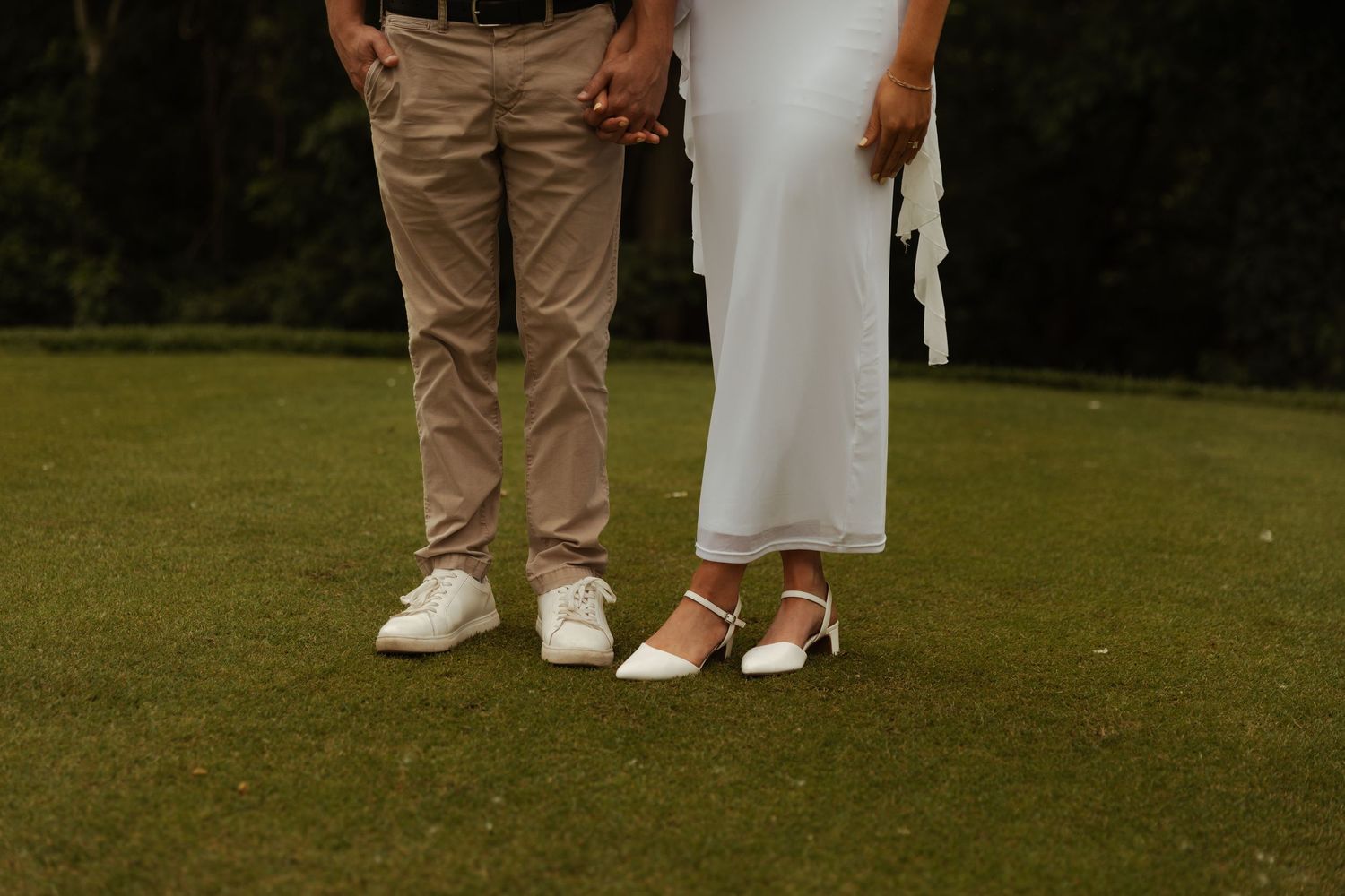 Bride in white dress and groom in khakis hold hands while standing on green grass.