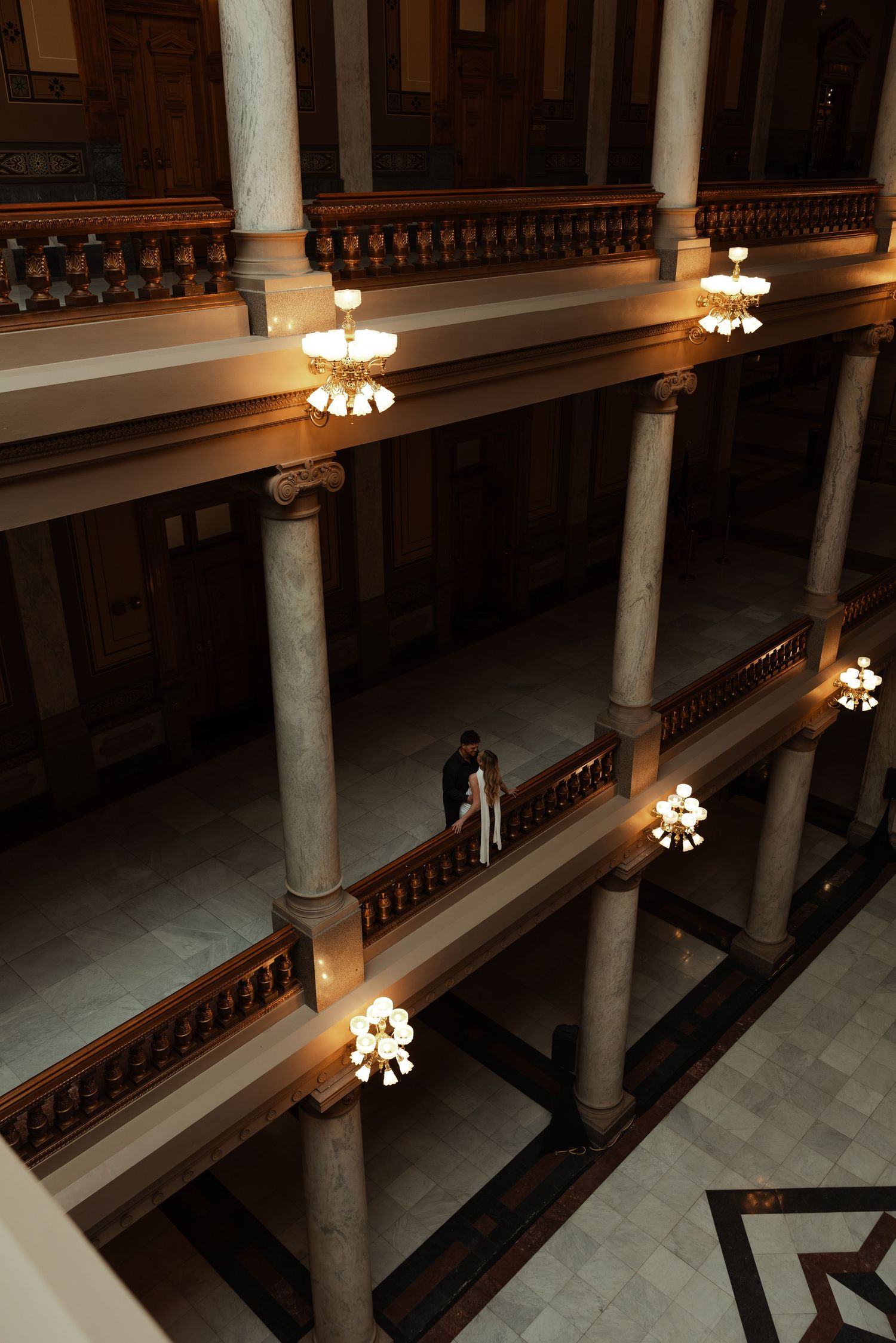 Aerial view of couple standing in grand historic building with ornate columns and chandeliers.