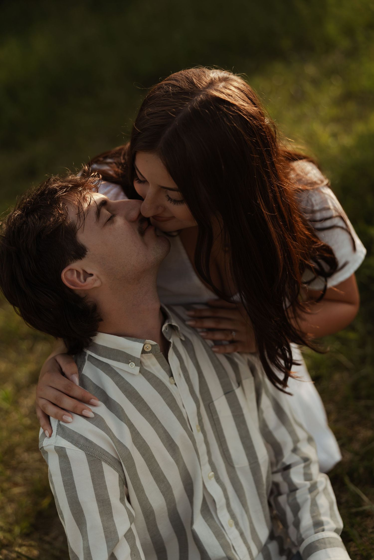 Romantic couple sharing a kiss outdoors in golden sunlight surrounded by lush green foliage.