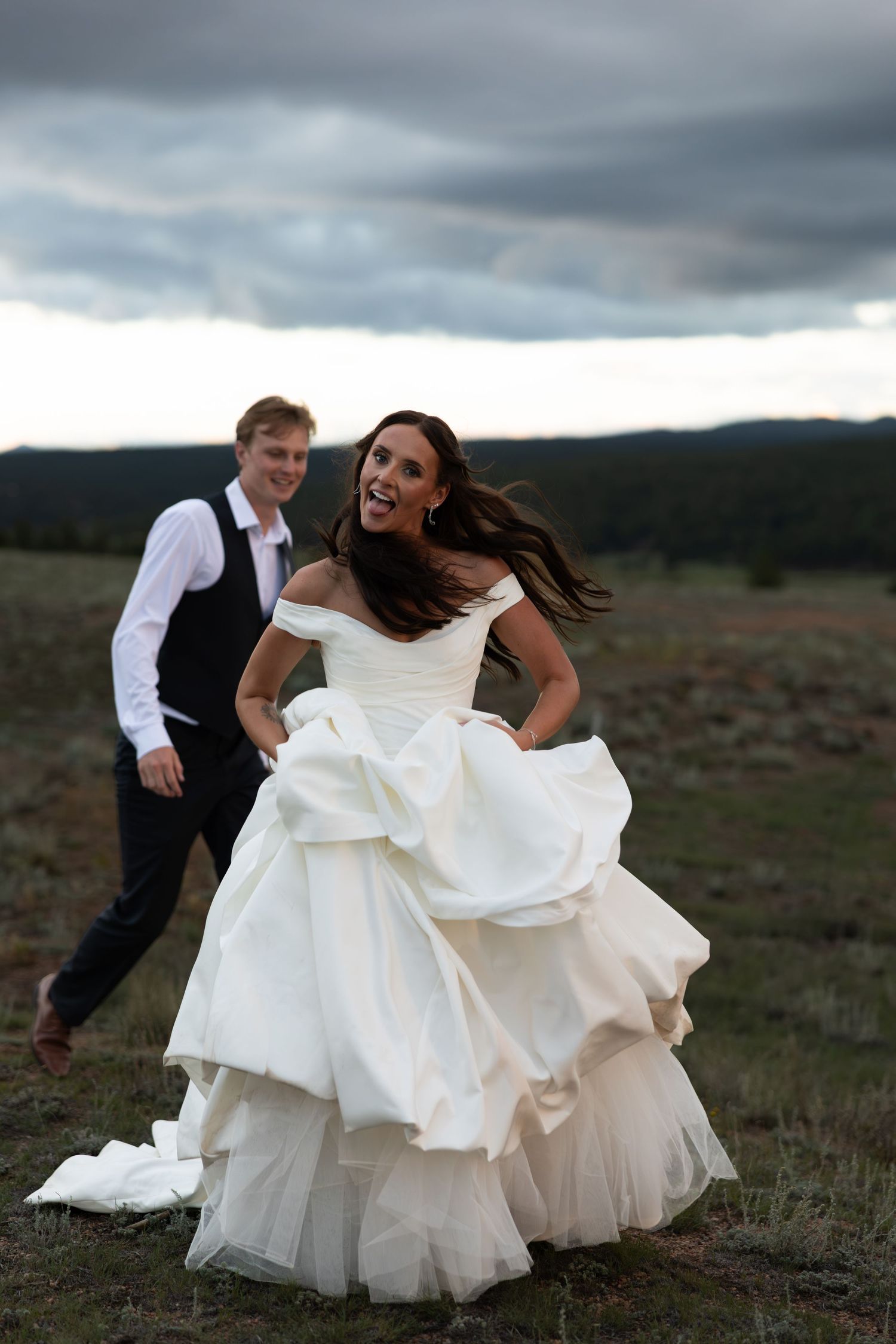 Joyful bride in layered white gown running with groom across open field under dramatic cloudy sky.