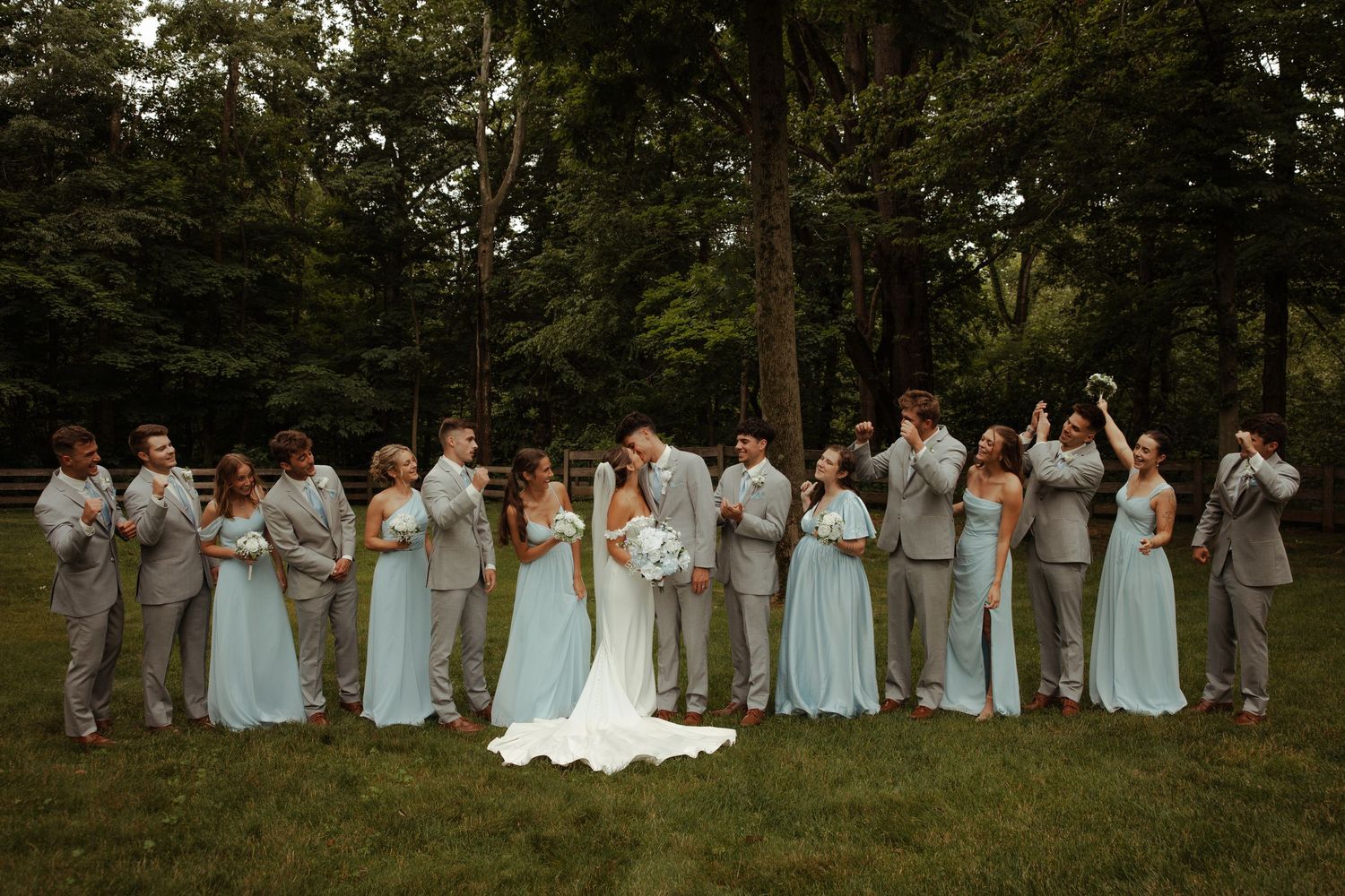 Large wedding party poses together on green lawn with bridesmaids in light blue gowns and groomsmen in gray suits.