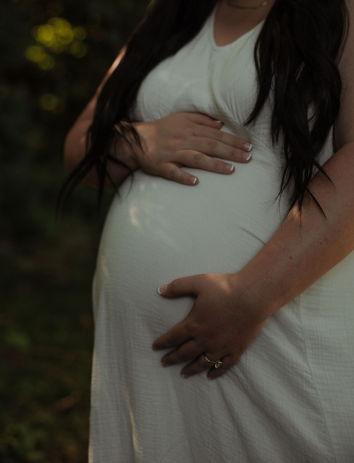 Close-up of pregnant woman in white dress gently holding baby bump with manicured nails and diamond ring.