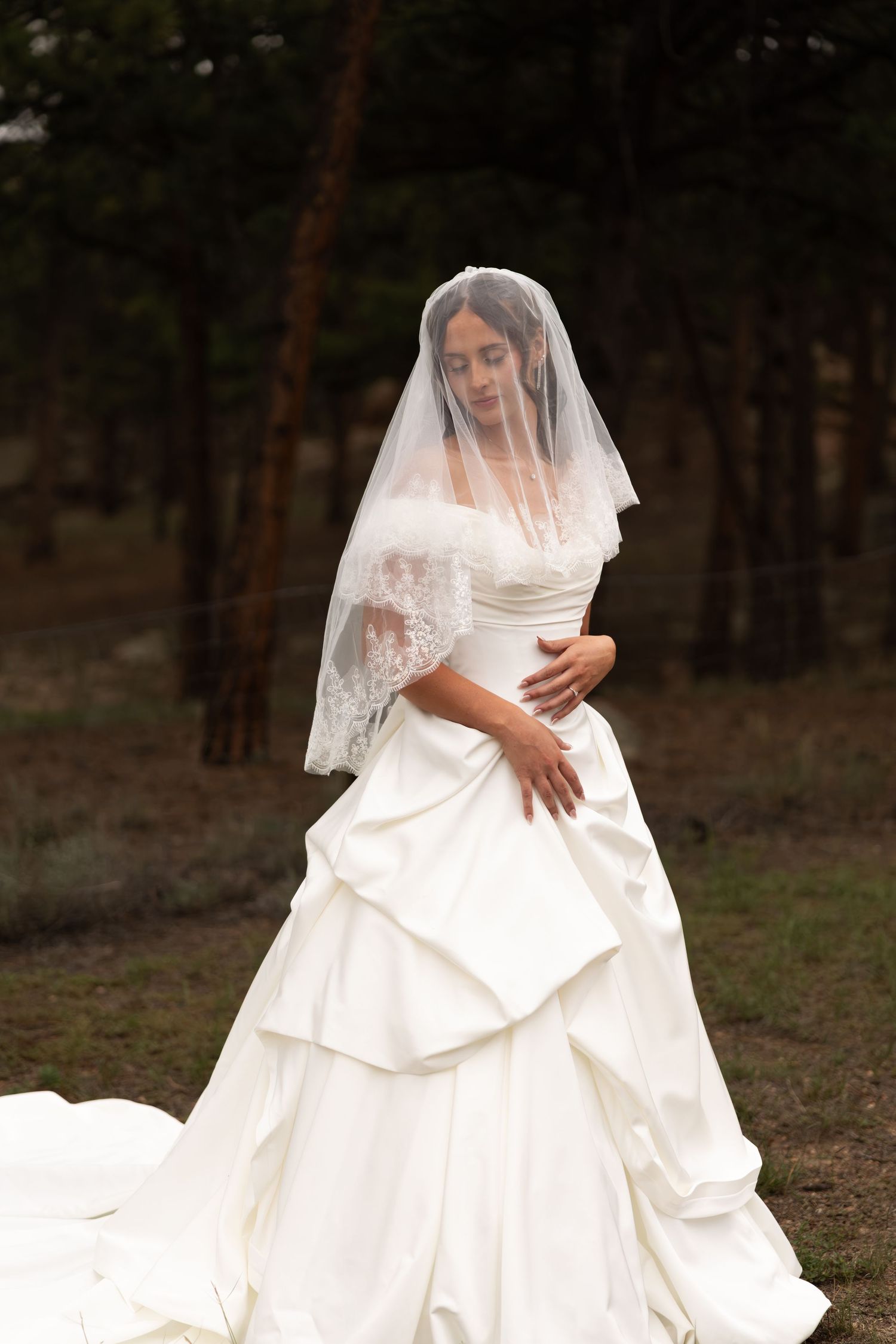 Bride in white ball gown and lace veil standing gracefully in a forest setting, looking down elegantly.