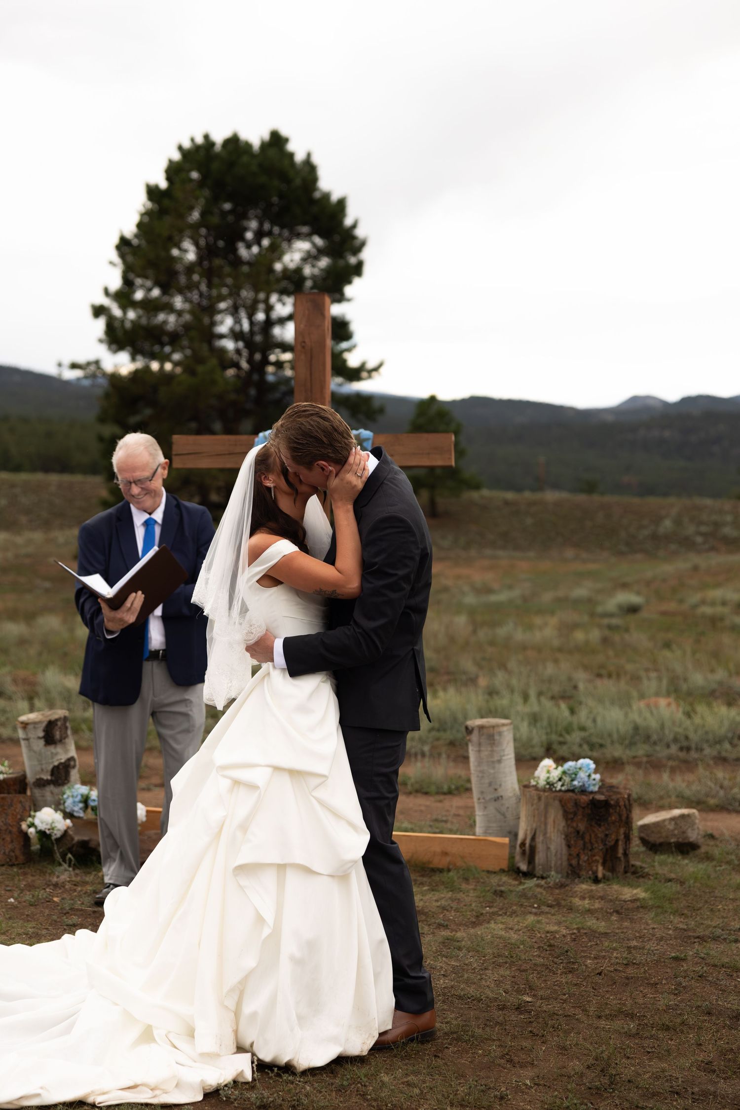 Bride and groom share first kiss at outdoor wedding ceremony with wooden cross and mountain backdrop.