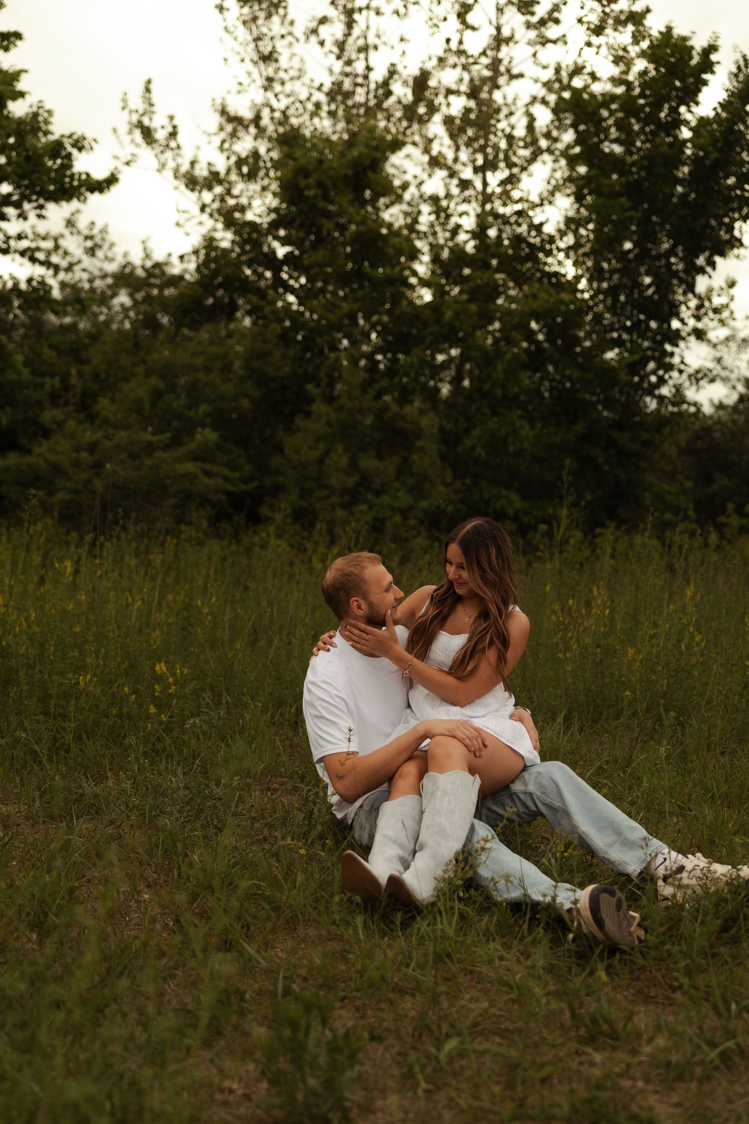 A couple sits together in a green meadow, sharing a romantic embrace during an outdoor engagement session.