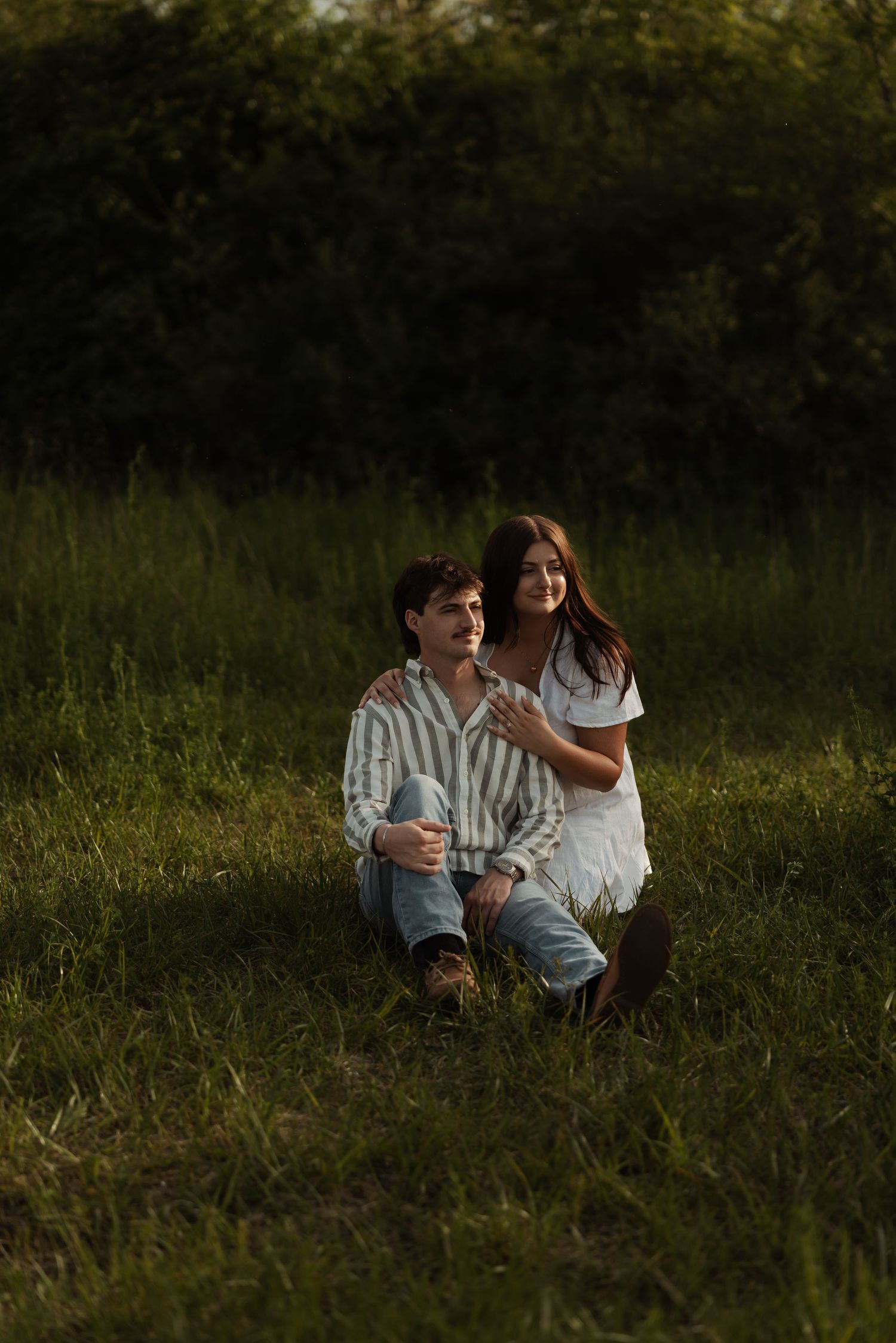 Couple sits together in golden hour sunlight on lush green grass during romantic outdoor engagement session.