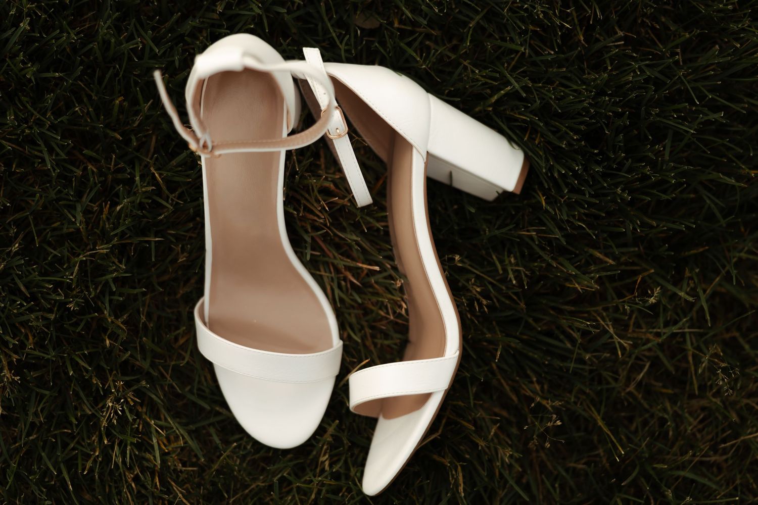 Elegant white block heel sandals with ankle strap resting on lush green grass for wedding.