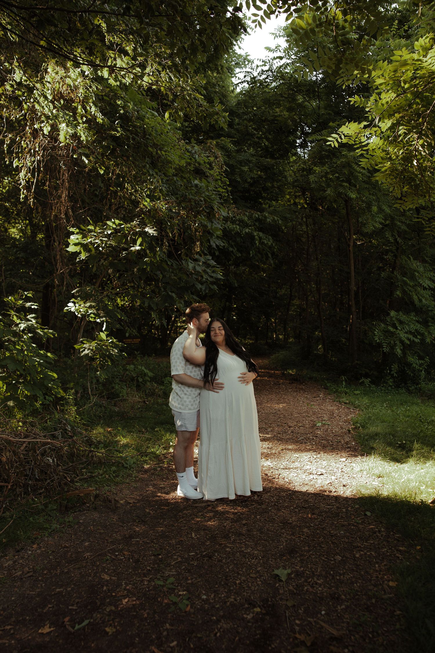 Pregnant woman in white dress embraced by partner on a sunlit forest path surrounded by lush green trees.