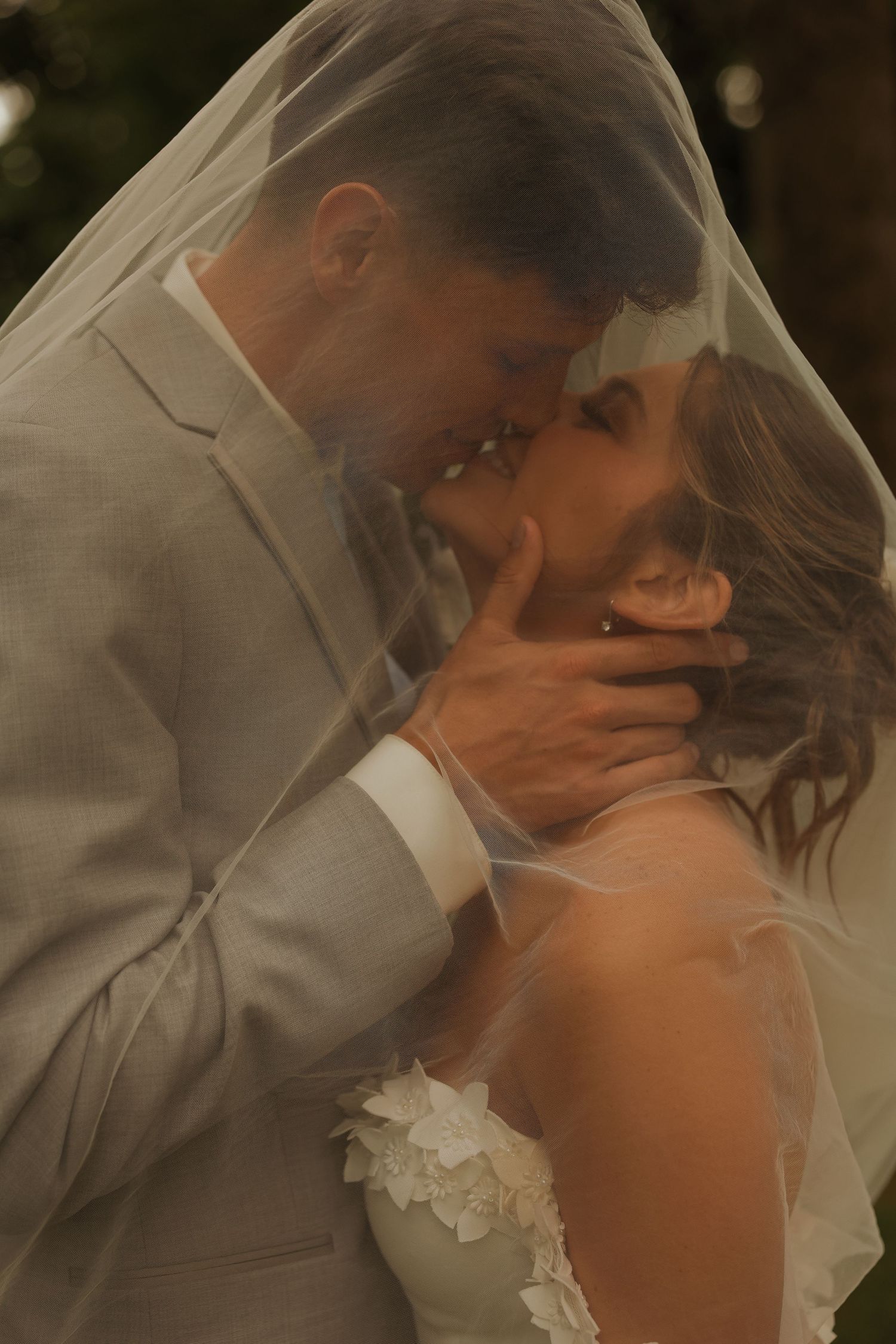 Bride and groom share passionate kiss under flowing wedding veil in romantic outdoor photo.