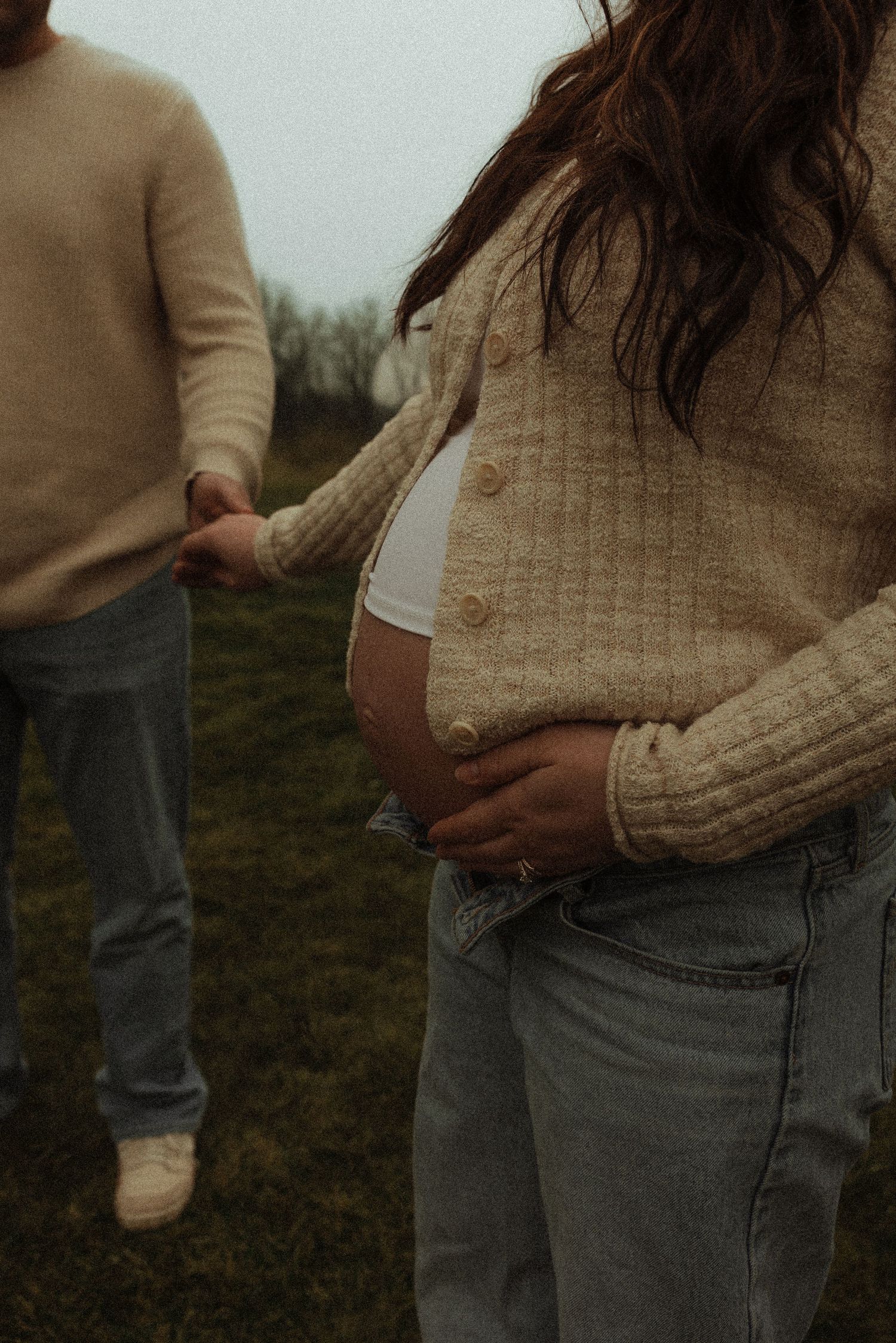 Pregnant woman holding baby bump while partner holds hand during outdoor maternity photoshoot.