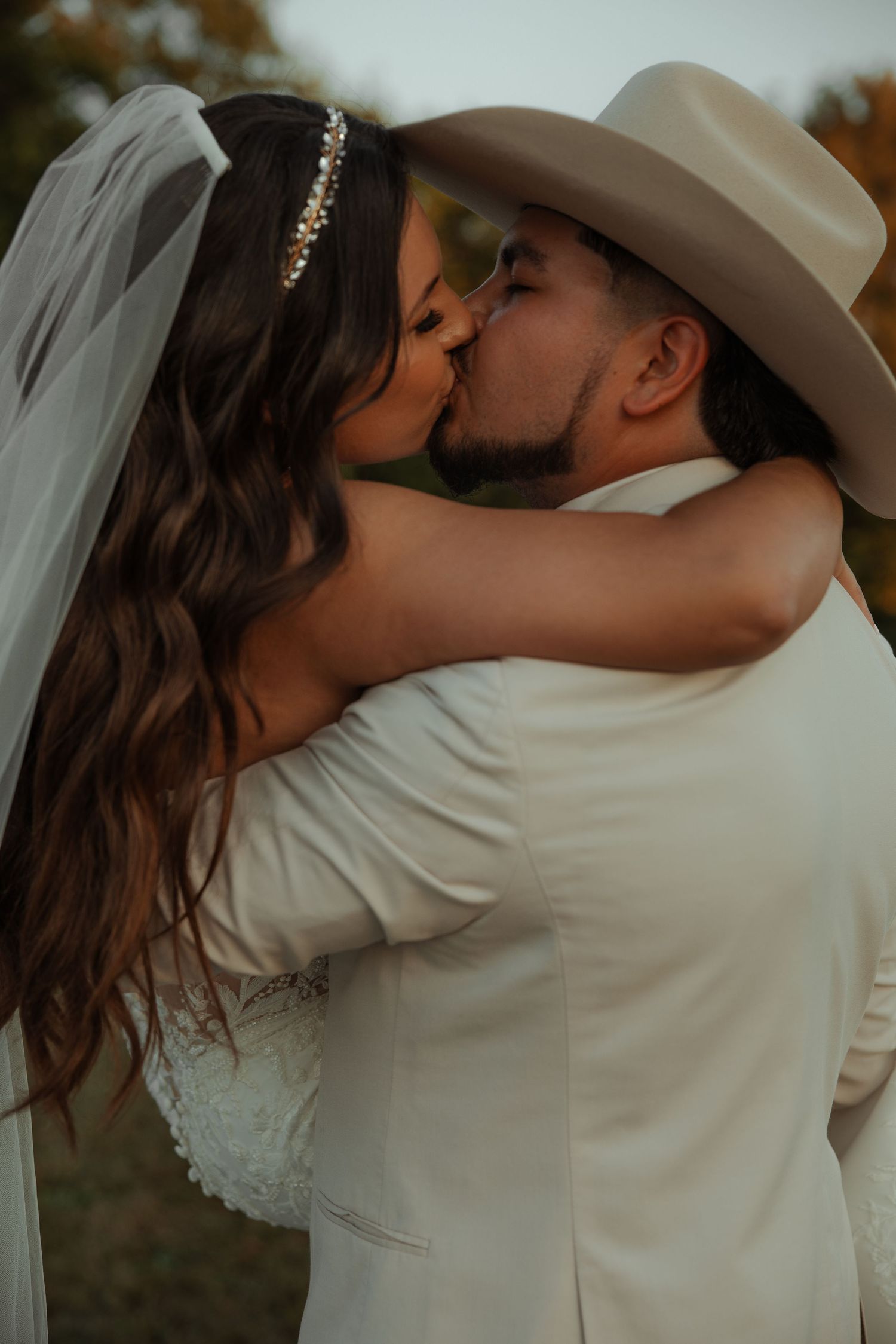 Bride with veil and groom in tan cowboy hat share passionate kiss outdoors in autumn.