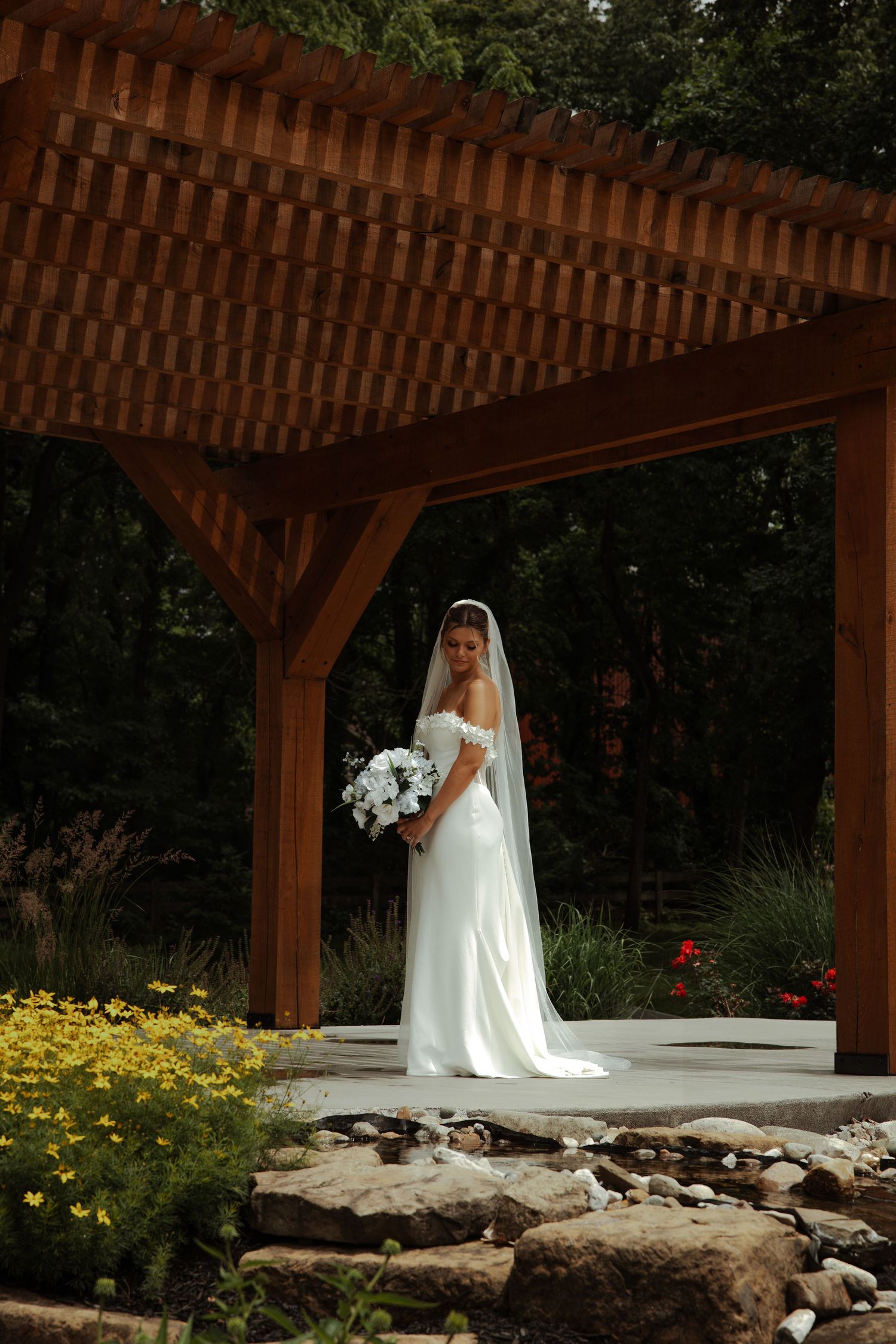 Elegant bride in off-shoulder white gown with veil poses under a wooden pergola in a garden setting.