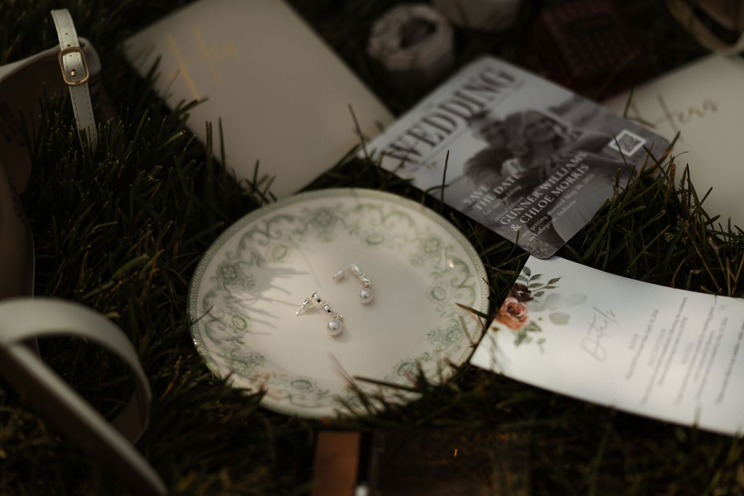 Wedding detail flat lay with earrings on decorative plate, program, and accessories on grass.