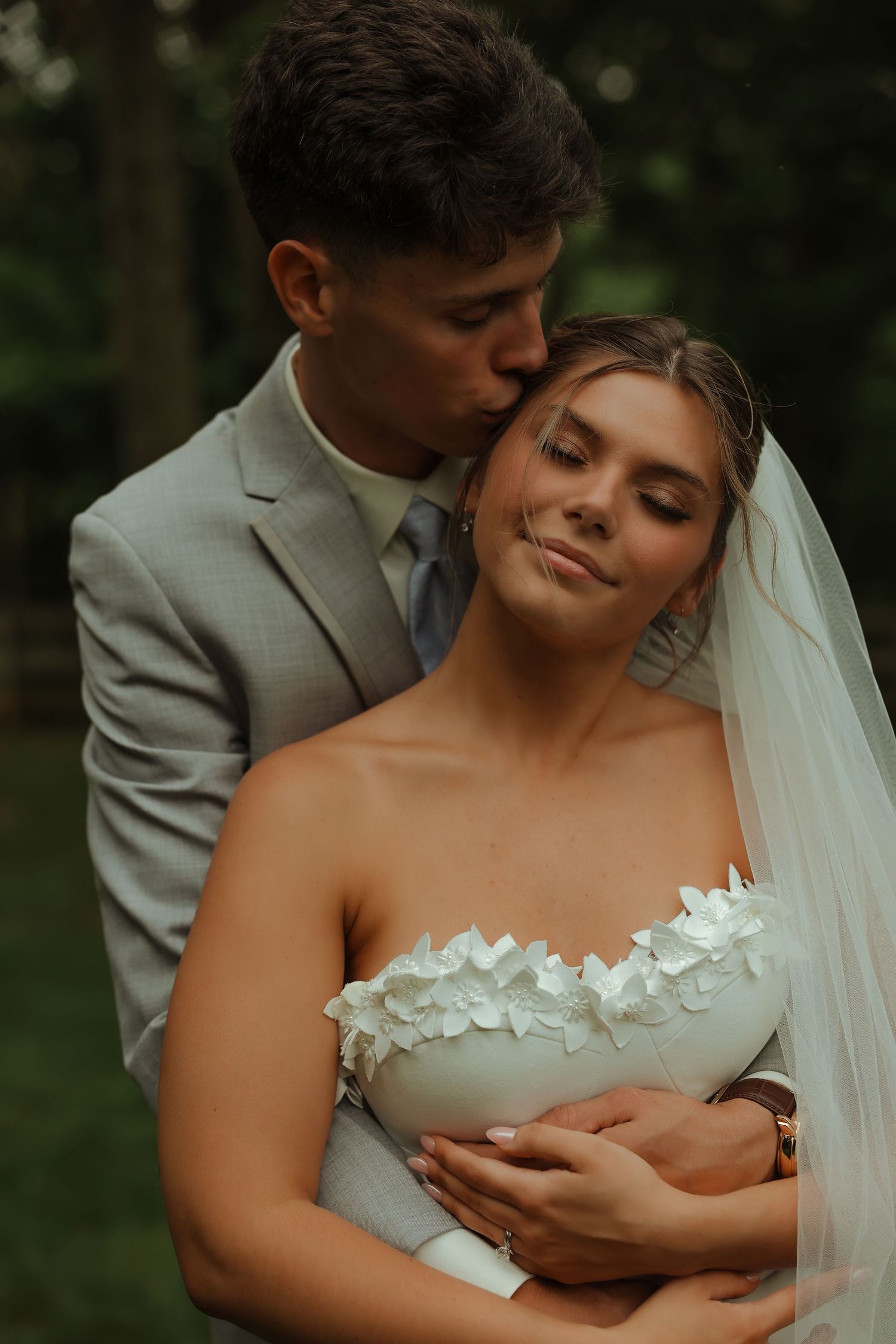 Groom in gray suit tenderly kissing bride on temple in lush green forest setting.