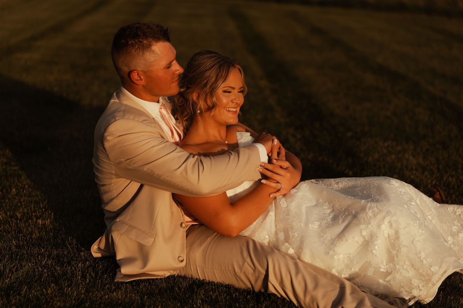 Bride and groom sitting on grass, laughing together in warm golden sunset light.