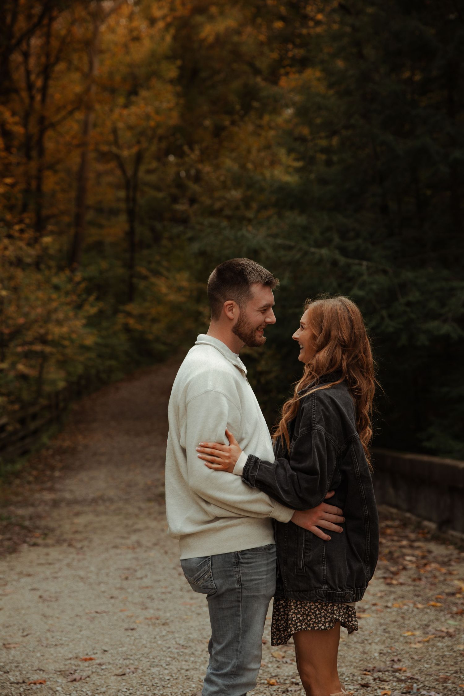 A smiling couple embraces on a gravel path surrounded by vibrant autumn foliage in a forest.