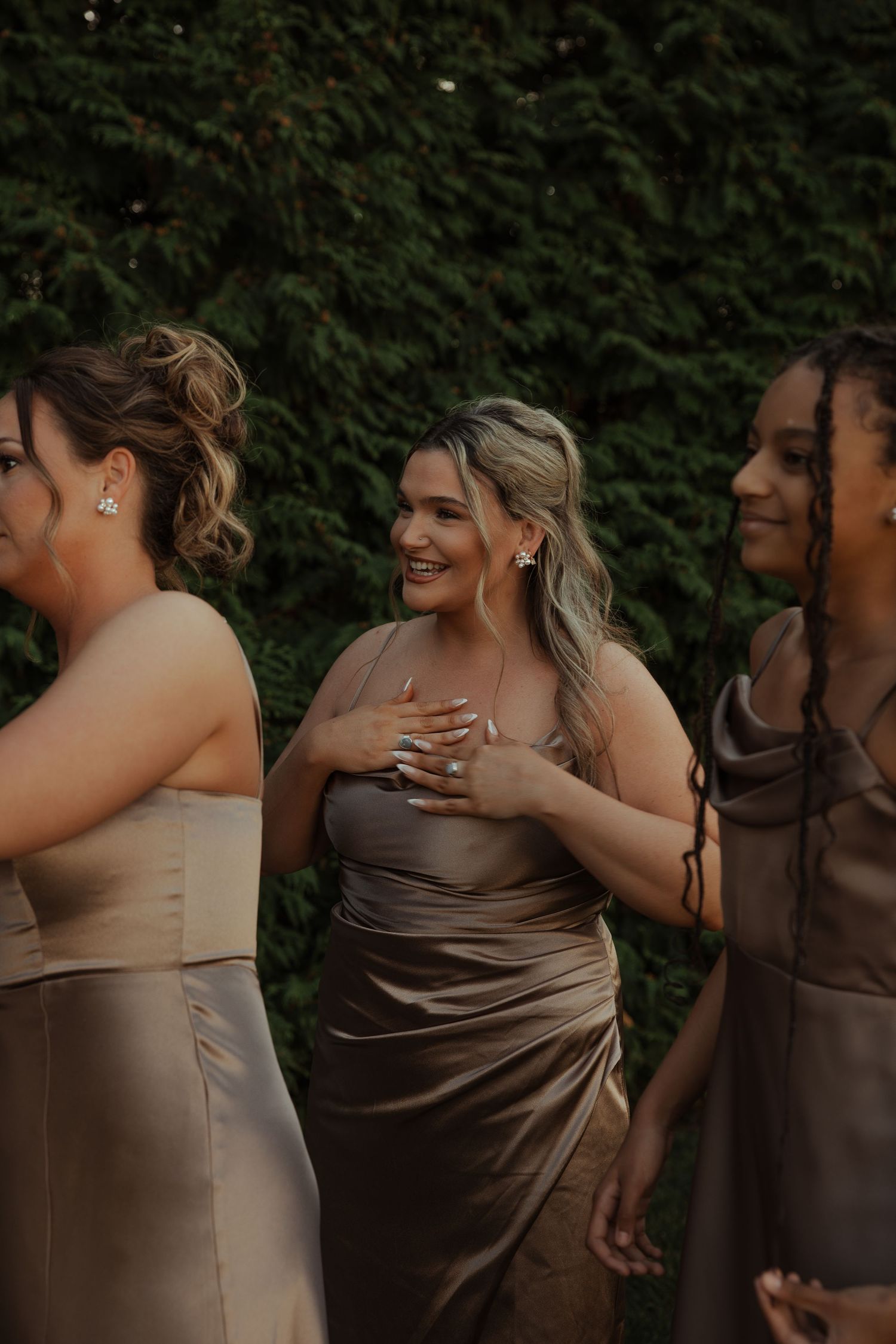 Three bridesmaids in elegant taupe satin dresses laughing and smiling together outdoors.