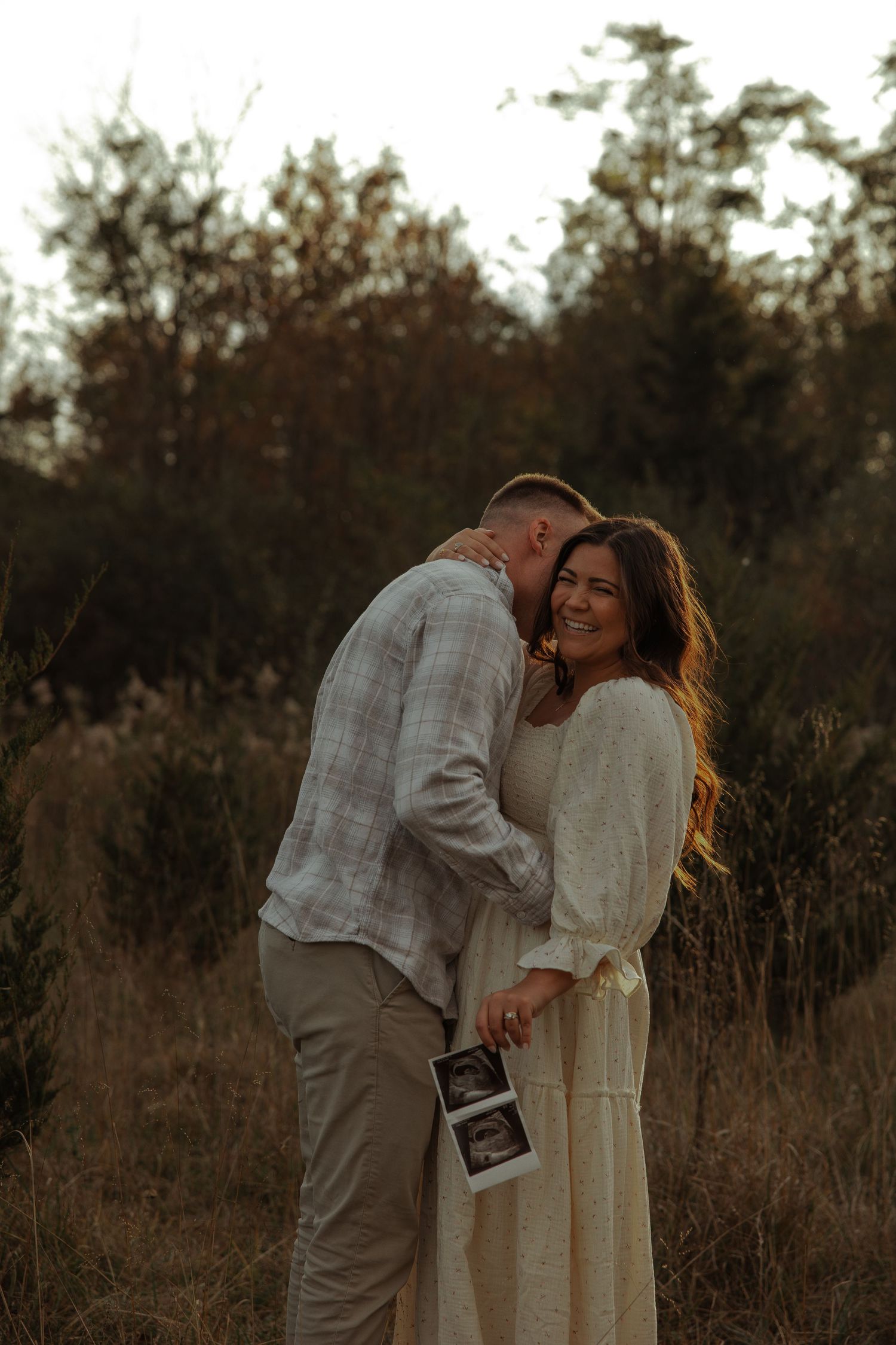 Couple embracing outdoors holding ultrasound photos during golden hour pregnancy announcement photoshoot.