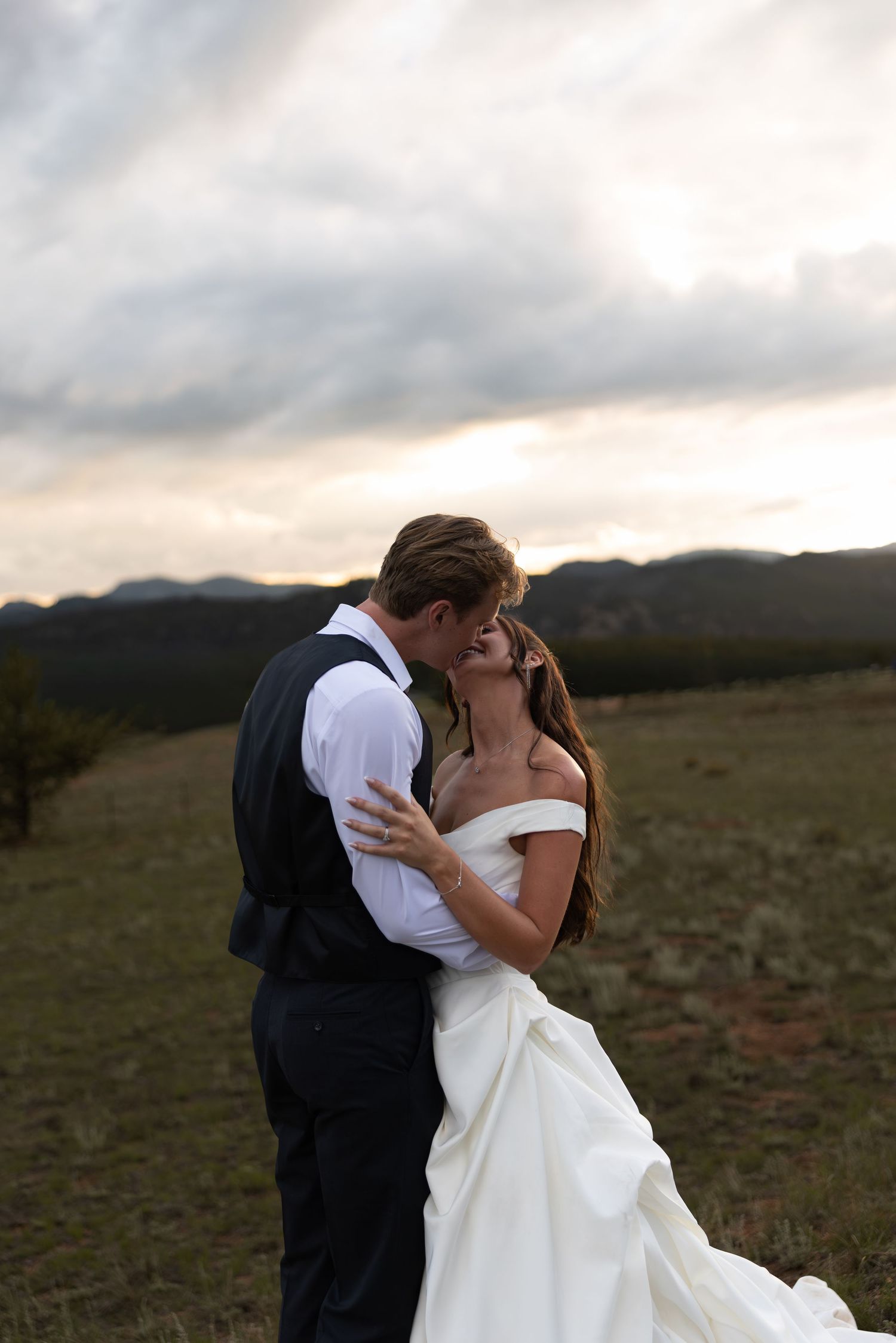 A bride and groom share a romantic embrace at sunset with mountain landscape backdrop on their wedding day.