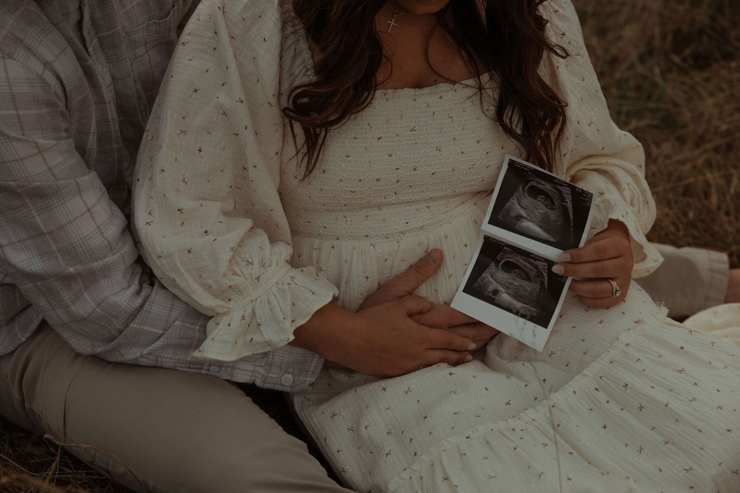 Close-up of pregnant couple holding ultrasound photos while sitting in a dry grass field.
