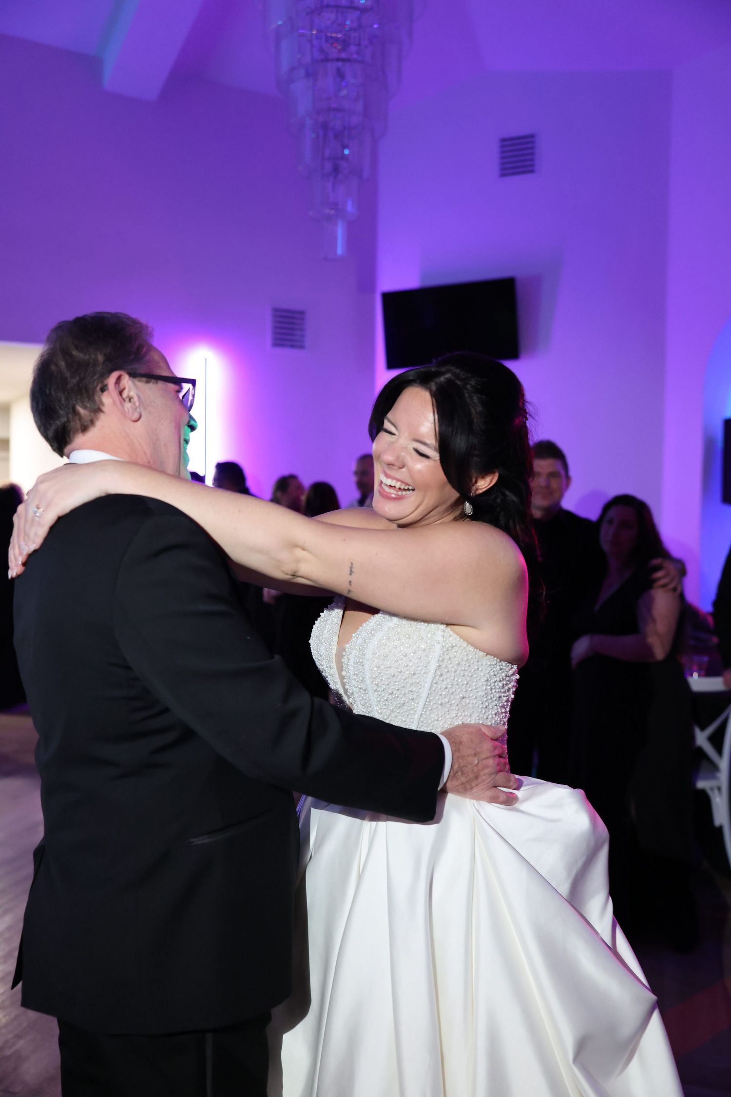 Bride in white satin gown smiling joyfully while dancing with father at wedding reception.