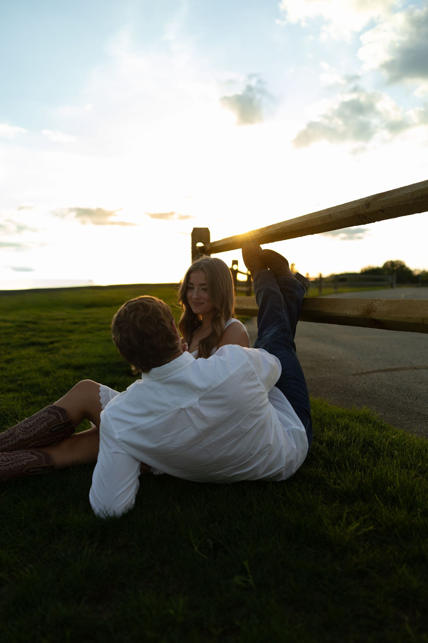 Couple sitting together on grass leaning against a wooden fence during golden hour sunset.