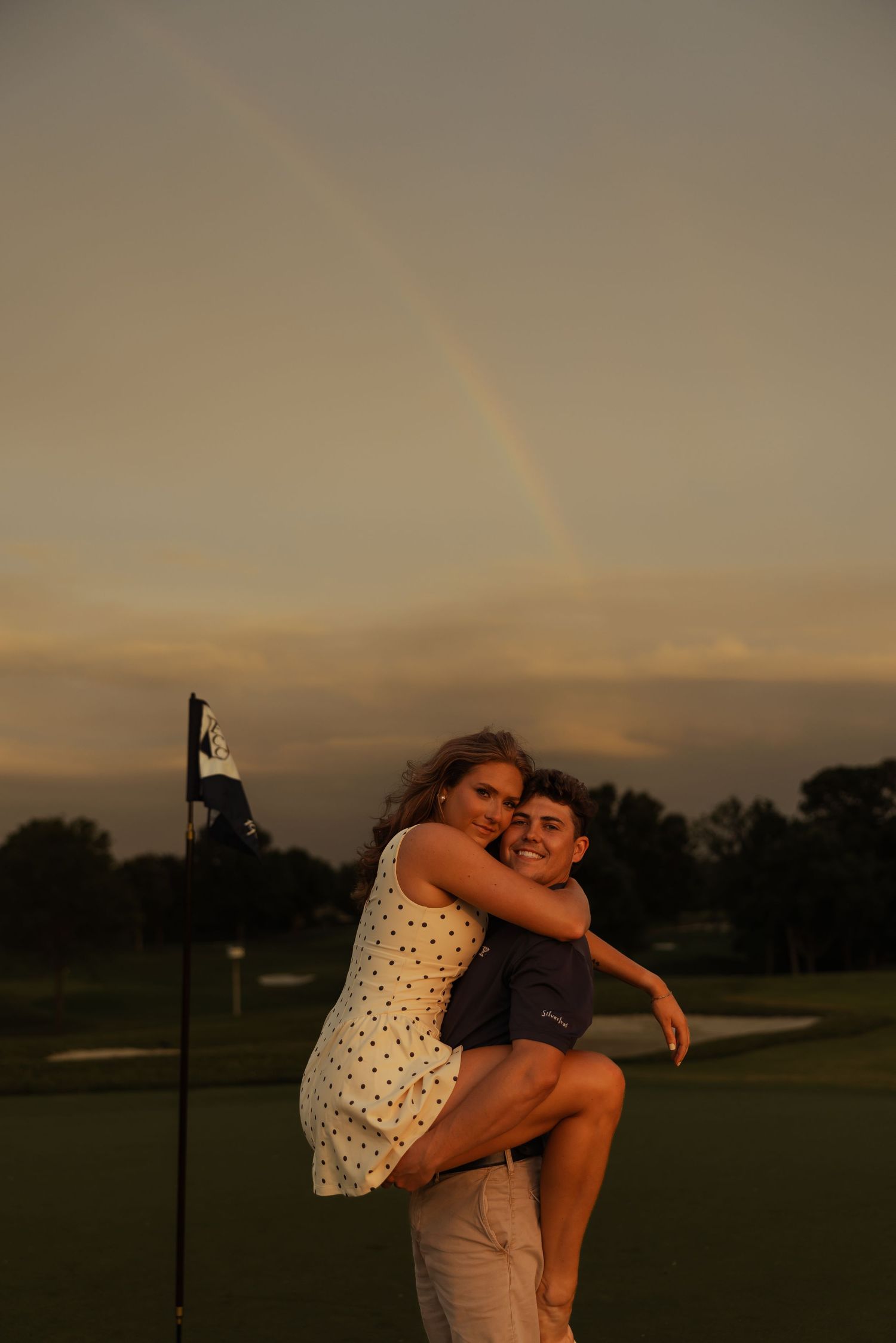 A couple embraces on a golf course at sunset with a rainbow visible in the sky behind them.