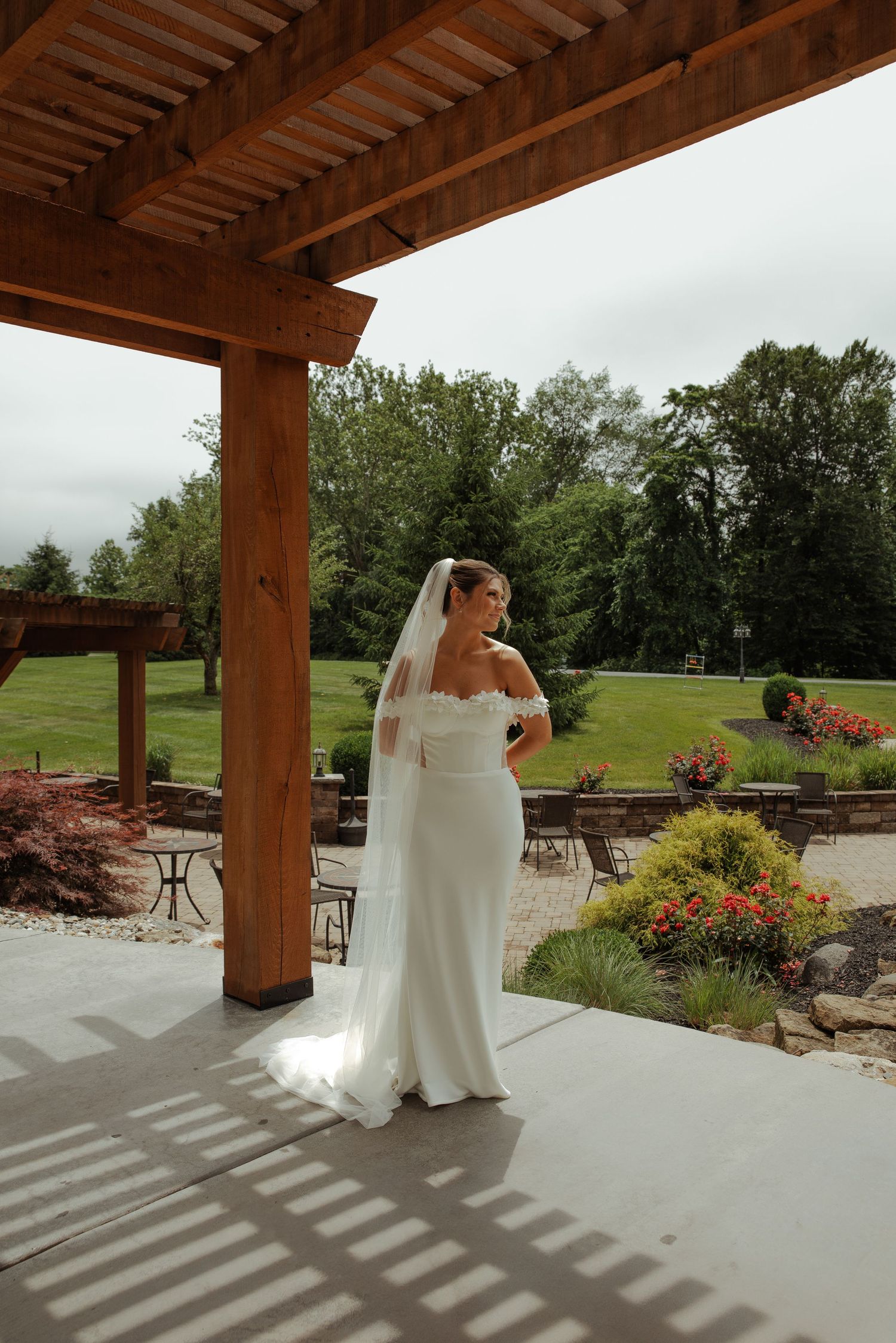 Elegant bride in a white strapless gown and veil standing under a wooden pergola at a garden venue.