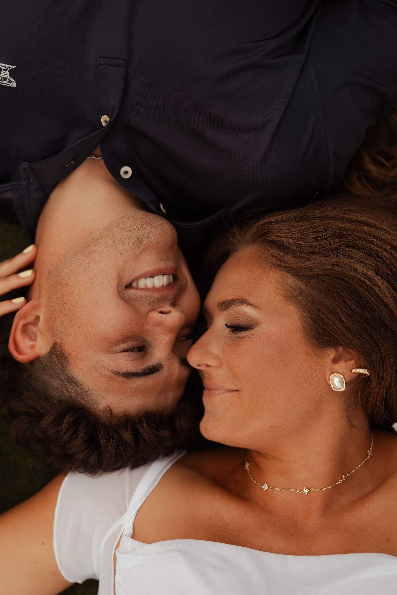 Smiling couple lies on grass in playful upside-down portrait during outdoor engagement photo session.