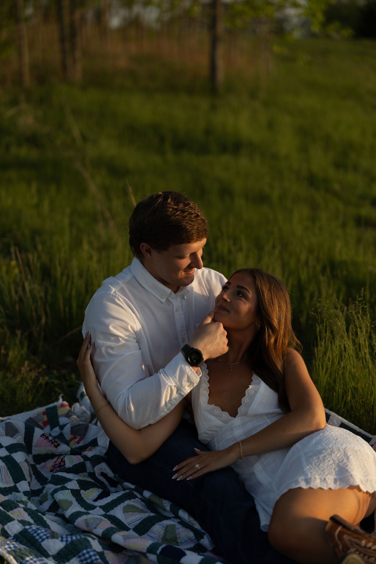 Engaged couple sitting on a quilt blanket in a green field during sunset.