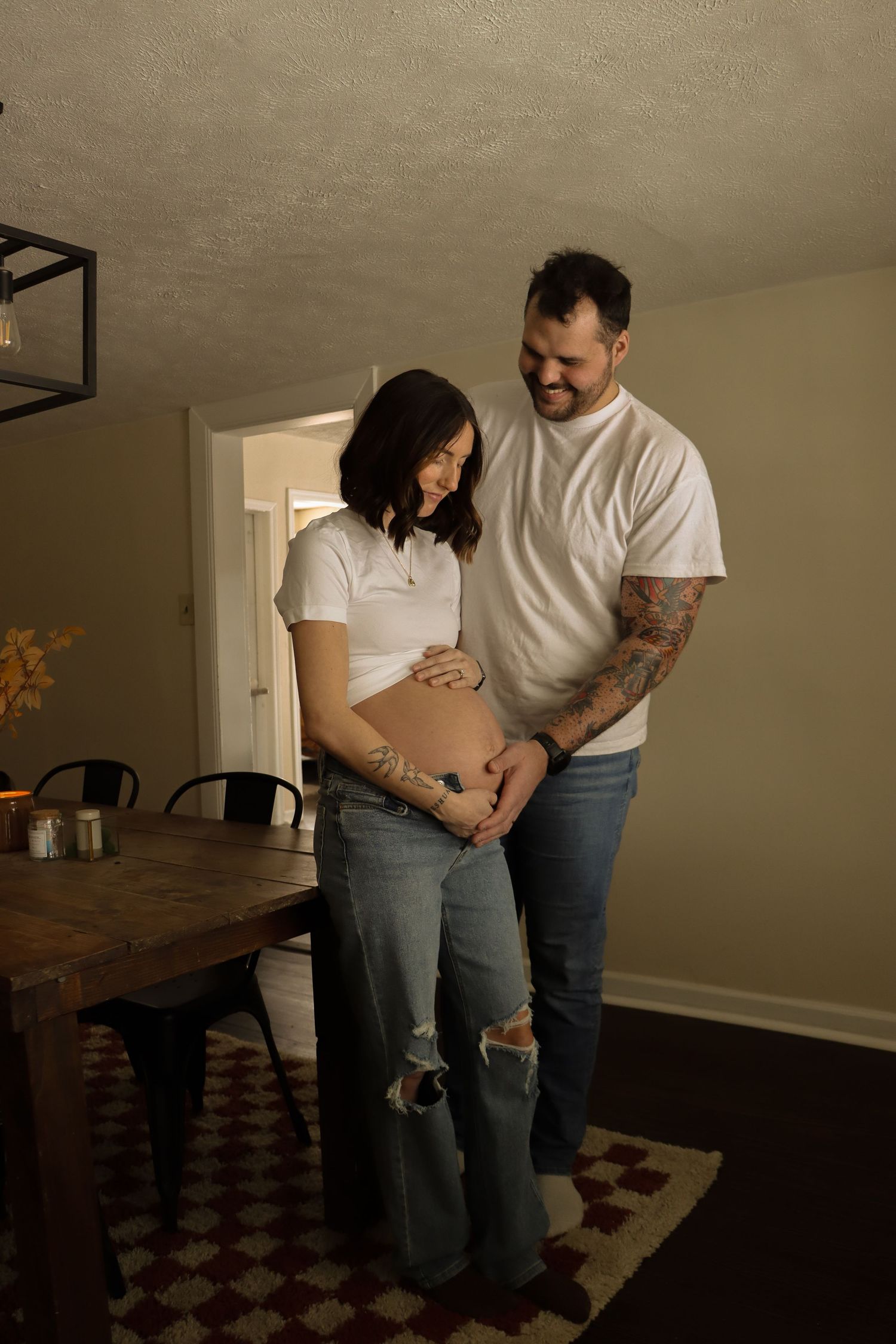 Smiling couple in white tops and jeans pose together showing pregnant belly in dining room.