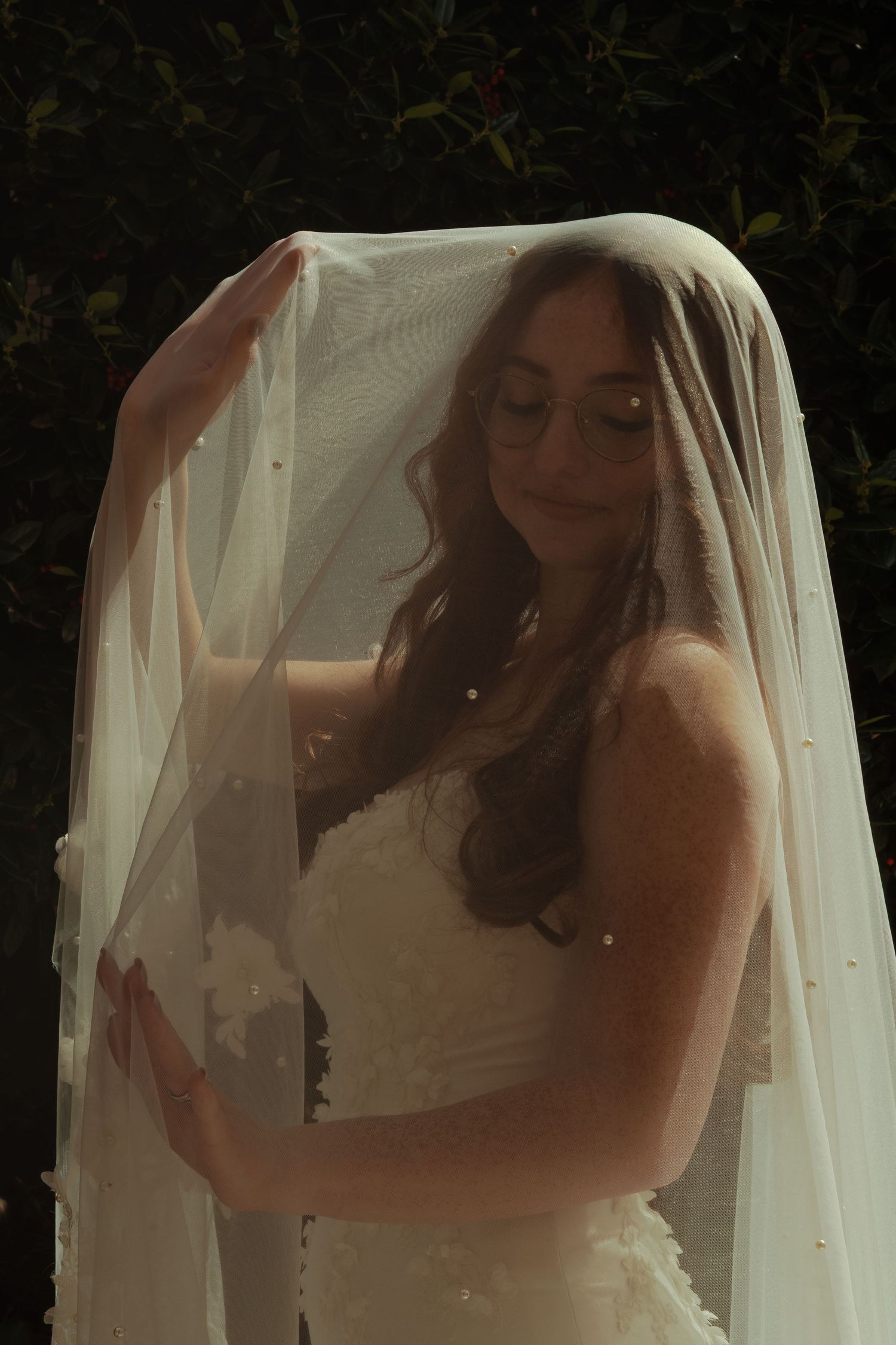 Bride in a white lace gown lifts her pearl-adorned veil, smiling softly in natural light.